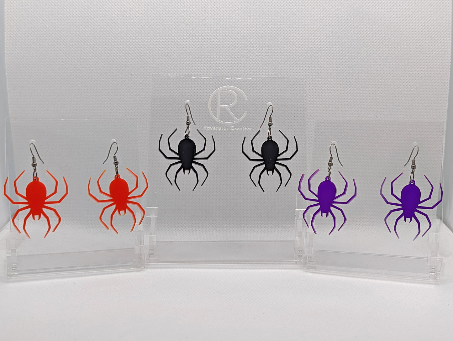Shows three pairs of earrings that have ear wire hook fixings. Each one is a spider with thin legs like a black widow with black fixings. The colours are red, black and purple. They are against a white background on see through stand.