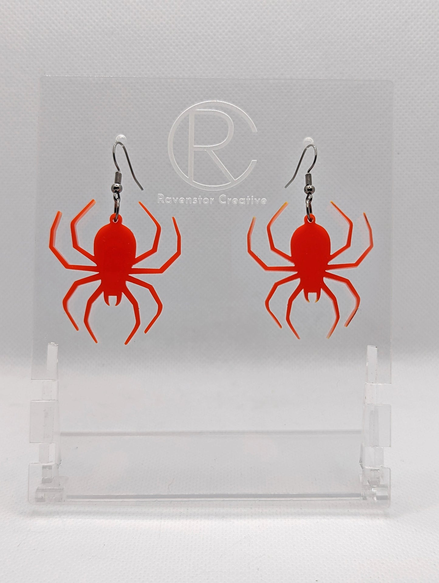 Shows a pair of earrings with black ear wire hook fixings. Each one is a spider with thin legs like a black widow. The colour of them is red. They are on a see through stand that has the Ravenstor Creative Logo and its against a white background.