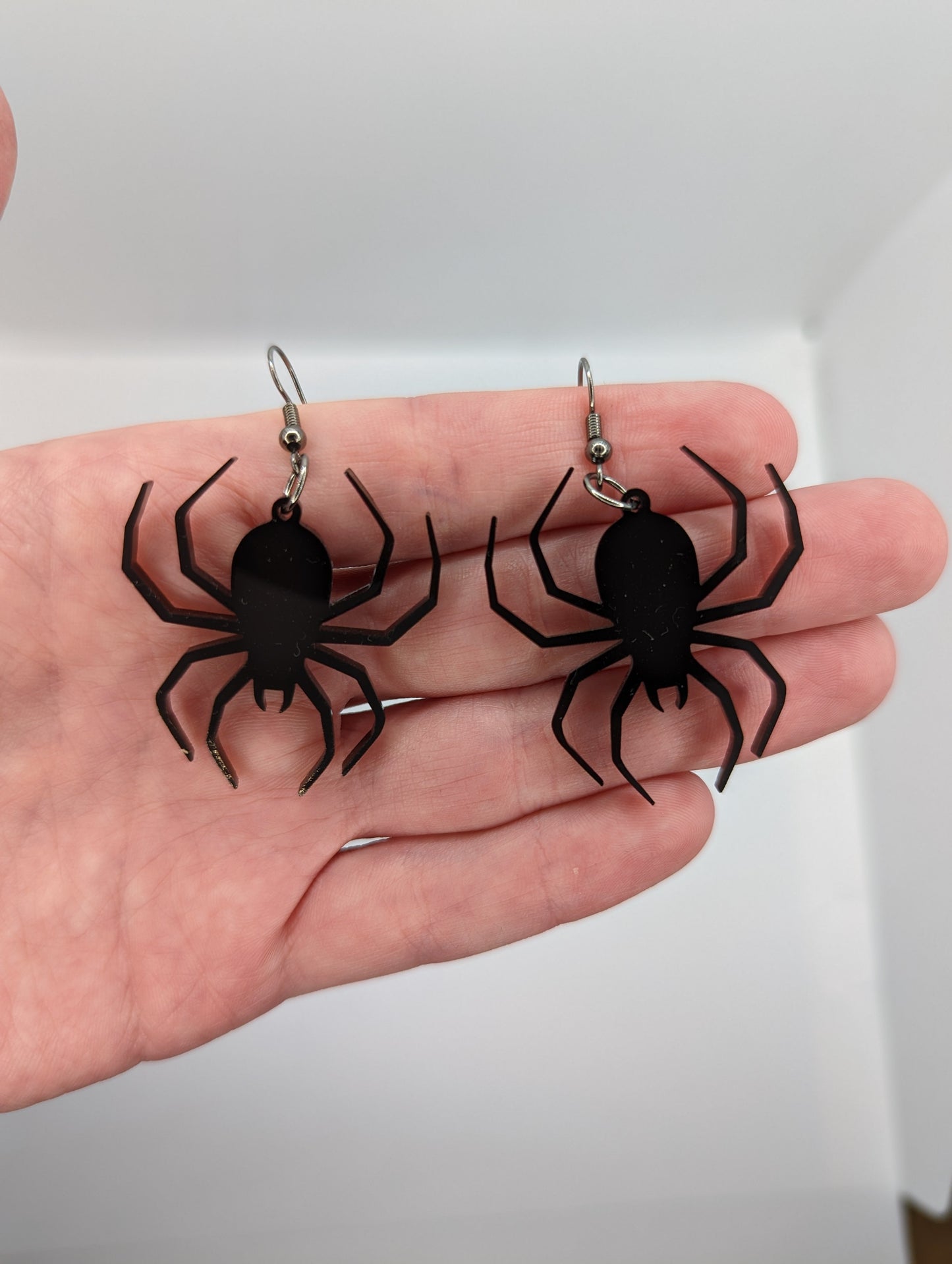 A pair of earrings in a hand to show the size with black fixings, each one is a spider with thin legs like a black widow. They are black in colour and hang like they are spinning a web, the earrings themselves are about 3 fingers width and length