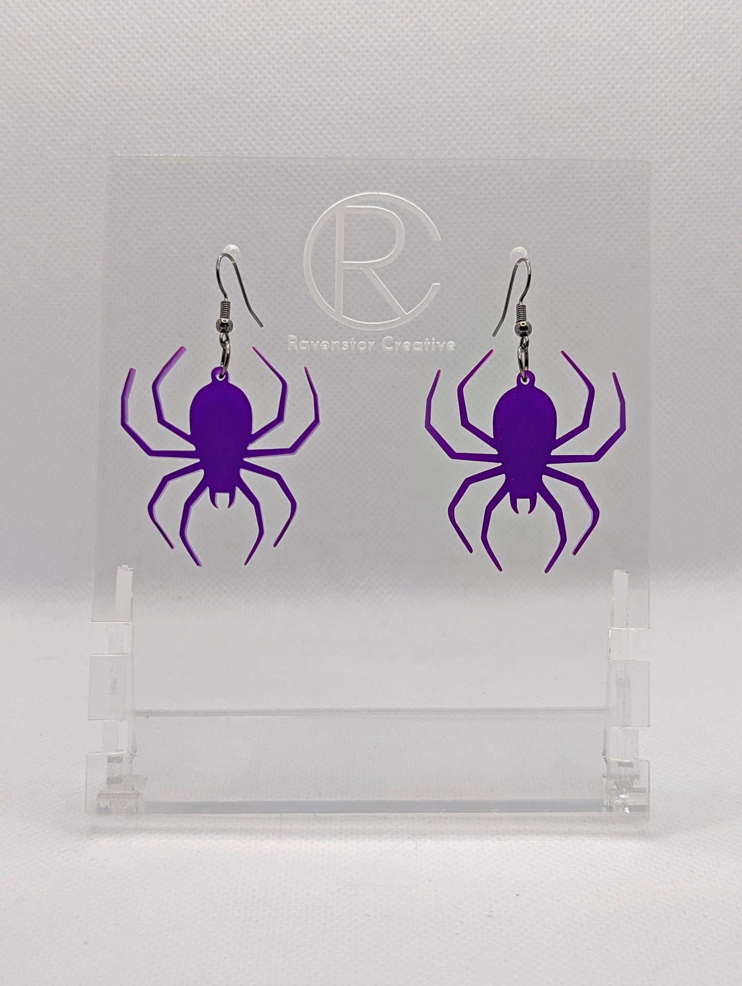 Shows a pair of earrings with black ear wire hook fixings. Each one is a spider with thin legs like a black widow. The colour of them is purple. They are on a see through stand that has the Ravenstor Creative Logo and its against a white background.