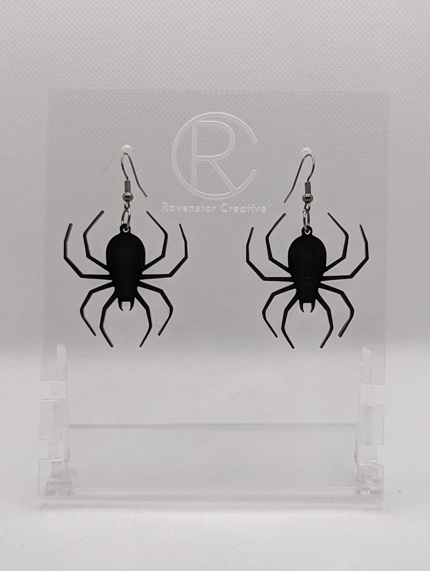 Shows a pair of earrings with black ear wire hook fixings. Each one is a spider with thin legs like a black widow. The colour of them is black. They are on a see through stand that has the Ravenstor Creative Logo and its against a white background.
