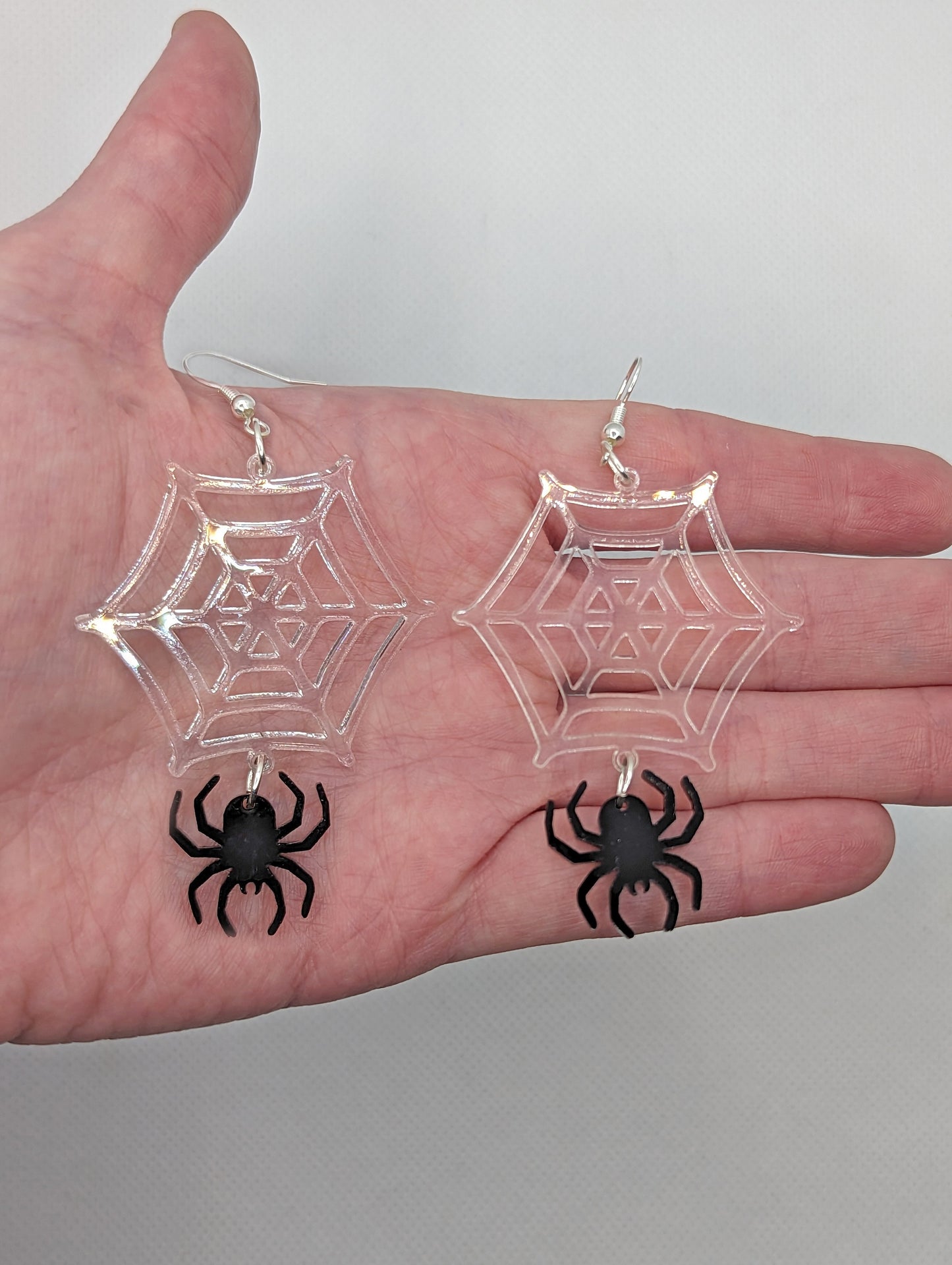 Image of a pair of spiders dangling from a web earrings on a hand showing the size against a white background, they are four finger widths in length and two and a half wide. The colours are a clear web and black spider and have silver fixings.