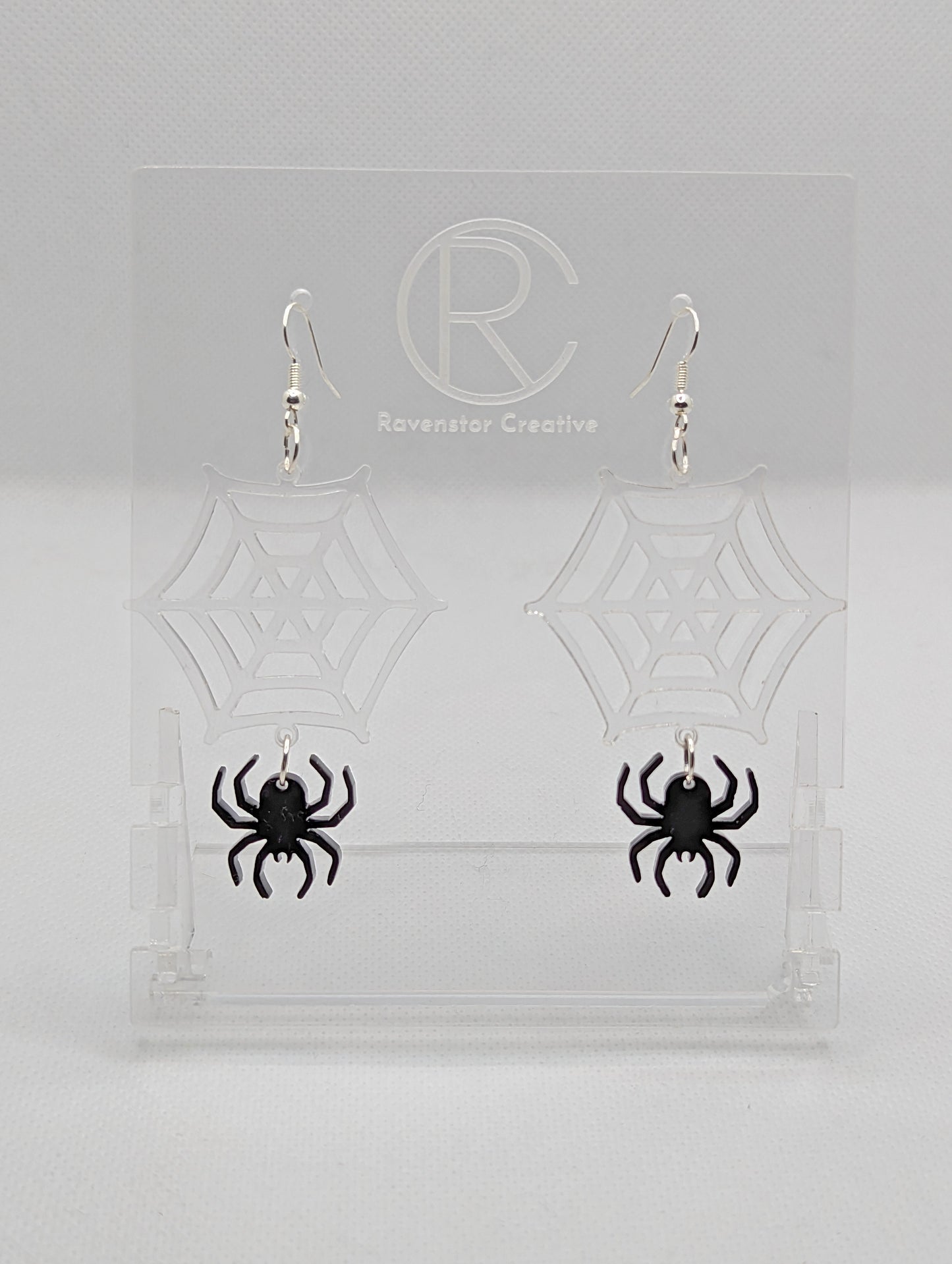 Shows a pair of earrings with silver ear wire hook fixings. They are spiders hanging from a web shapes. The colour of them are clear - see through for the web and black for the spider. They are on a see through stand against a white background.