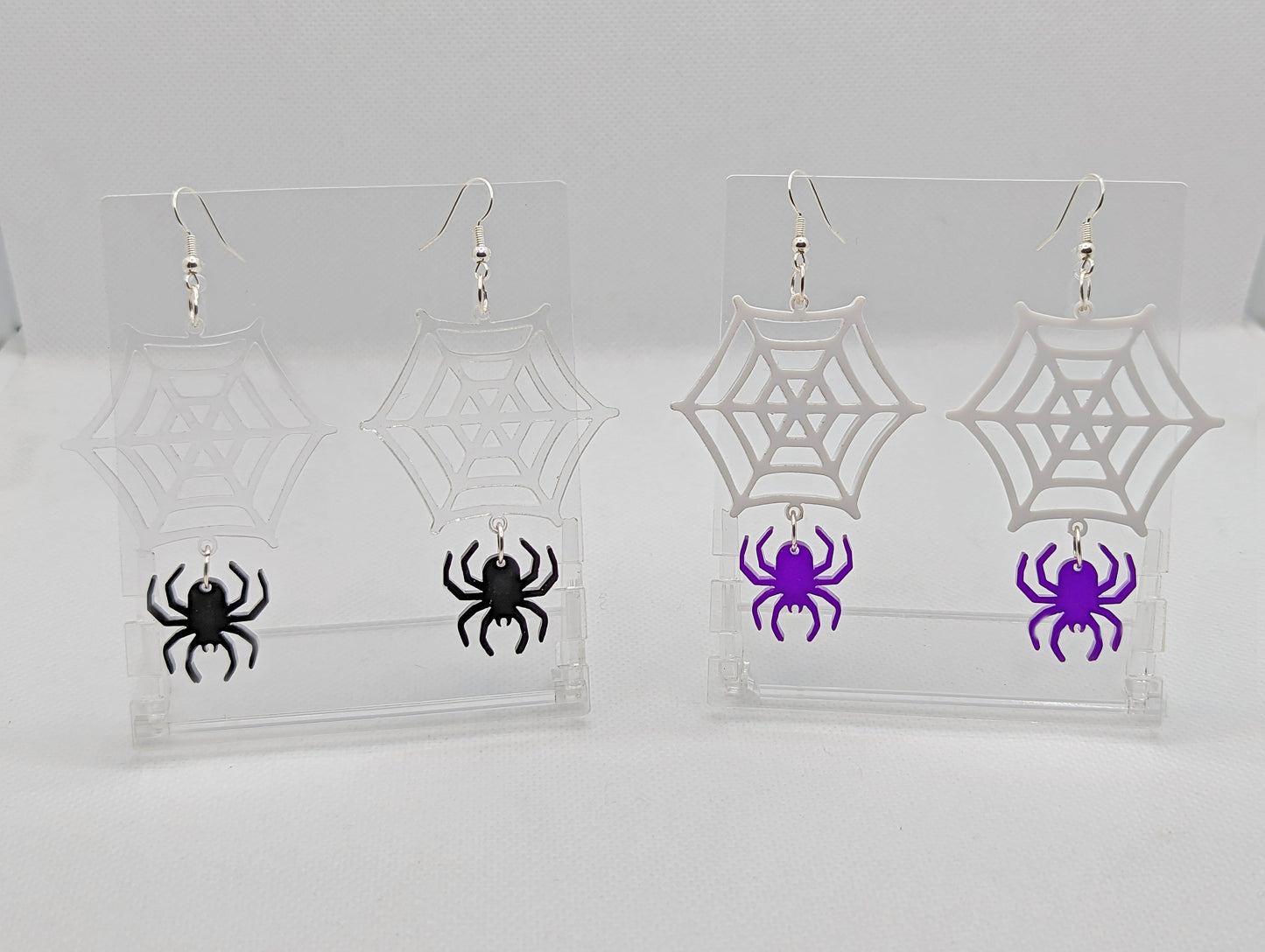 Shows 2 pairs of earrings with silver ear wire hook fixings. They are spiders dangling from a web. The colours of them are White web & purple spider and clear web & black spider. They are on see through stands against a white background.