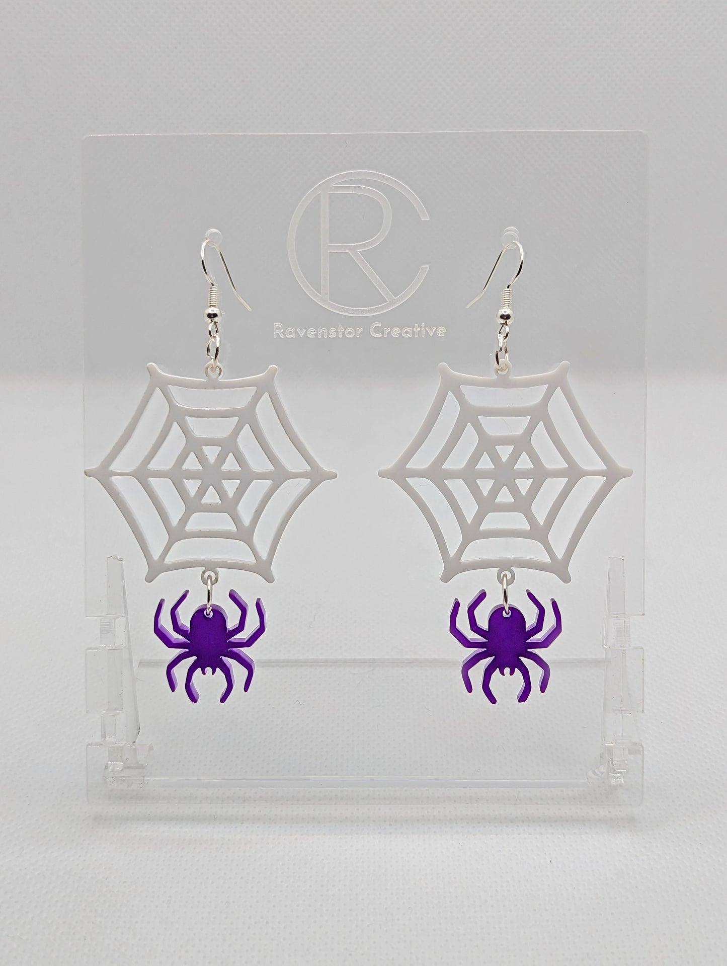 Shows a pair of earrings with silver ear wire hook fixings. They are spiders hanging from a web shapes. The colour of them are white for the web and purple for the spider. They are on a see through stand against a white background.