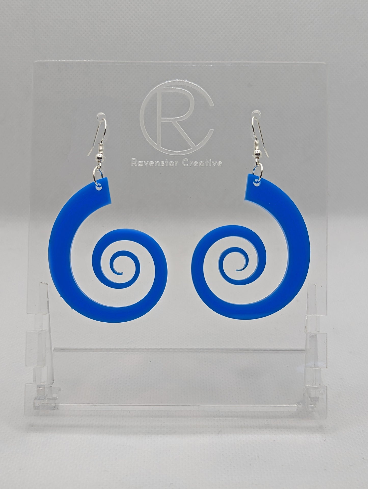 A pair of earrings with silver Ear wire hook fixings. They are large Retro 80's inspired spirals. They are made from blue acrylic. They are against a white background on a clear stand with the Ravenstor Creative logo on.