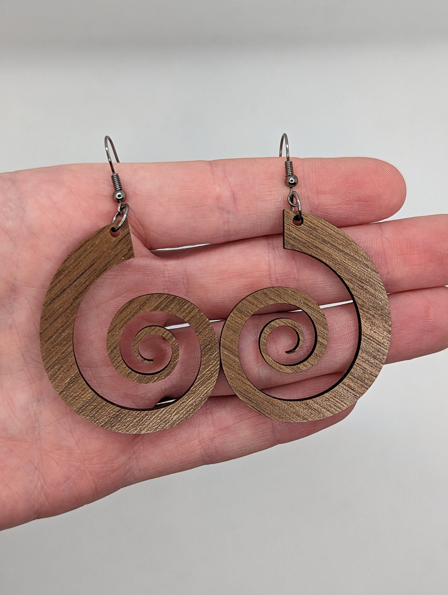 A pair of earrings against a hand showing the size. Retro 80's inspired spiral earrings with black ear wire hook fixings. They are 3 finger widths in length and same again wide. They are made from Walnut hardwood.