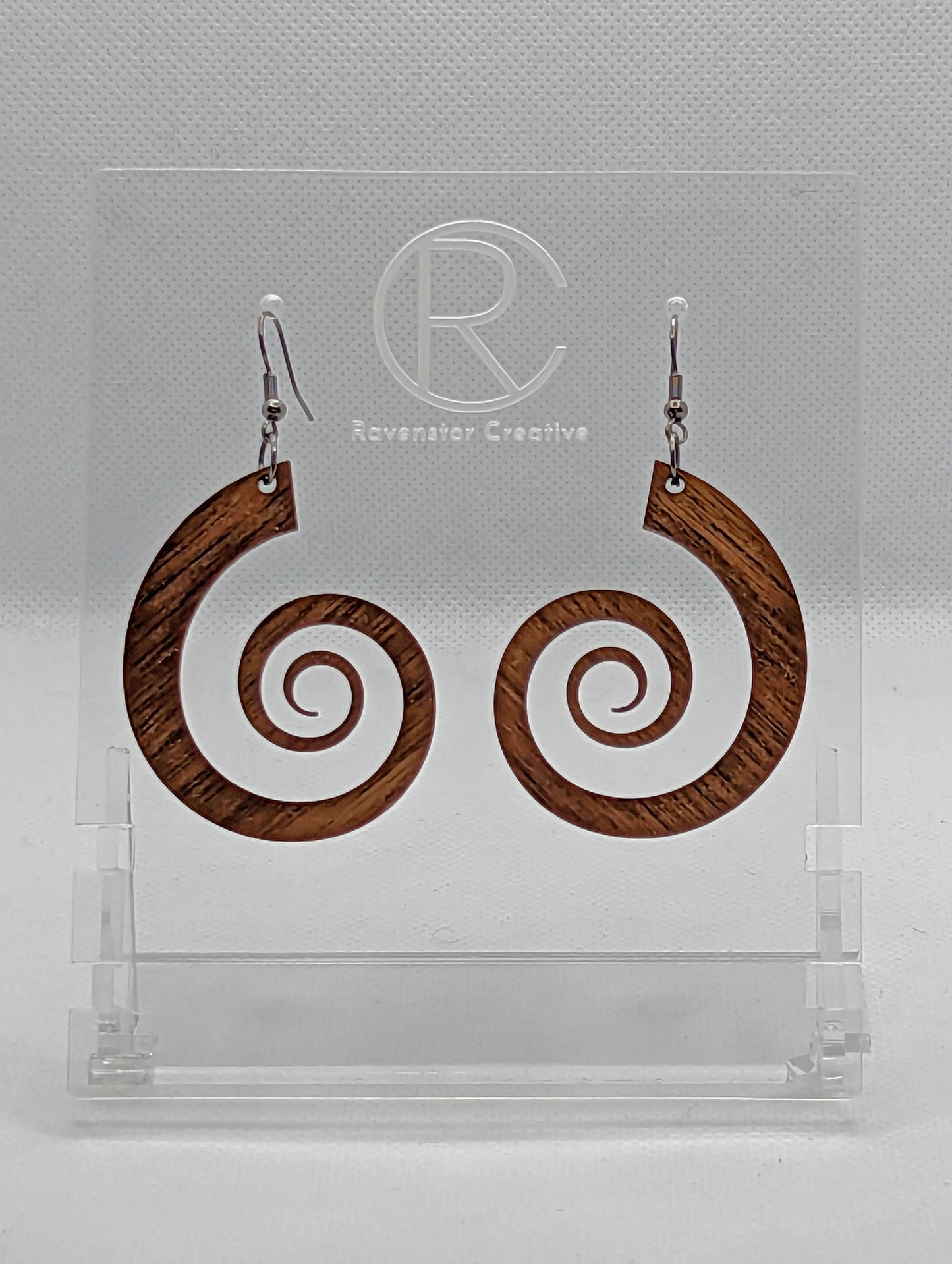 A pair of earrings with black Ear wire hook fixings. They are large Retro 80's inspired spirals. They are made from Walnut Wood. They are against a white background on a clear stand with the Ravenstor Creative logo on.