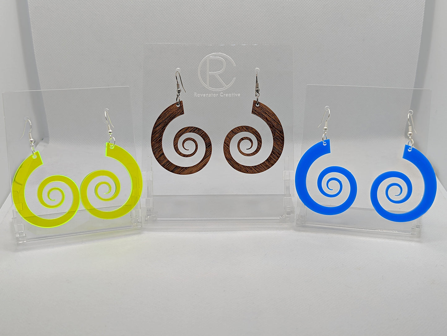 Three pairs of large Retro 80's inspired spiral earrings with Ear wire hook fixings. They are made from Neon Yellow acrylic, walnut wood and blue acrylic. They are on clear stands against a white background.