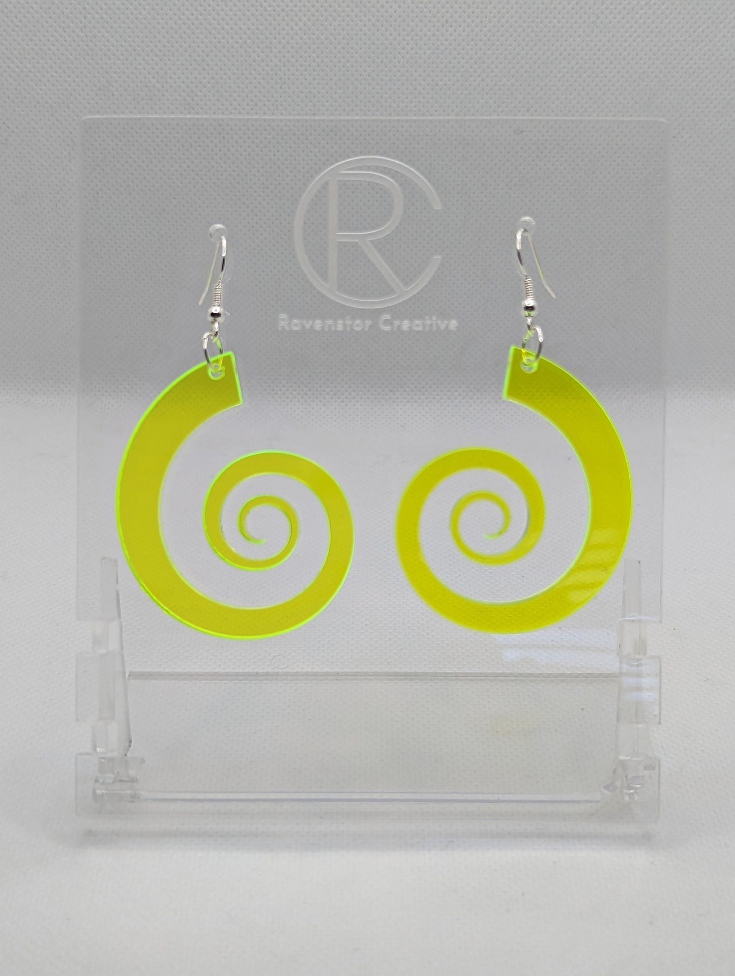 A pair of earrings with silver Ear wire hook fixings. They are large Retro 80's inspired spirals. They are made from Neon Green acrylic. They are against a white background on a clear stand with the Ravenstor Creative logo on.