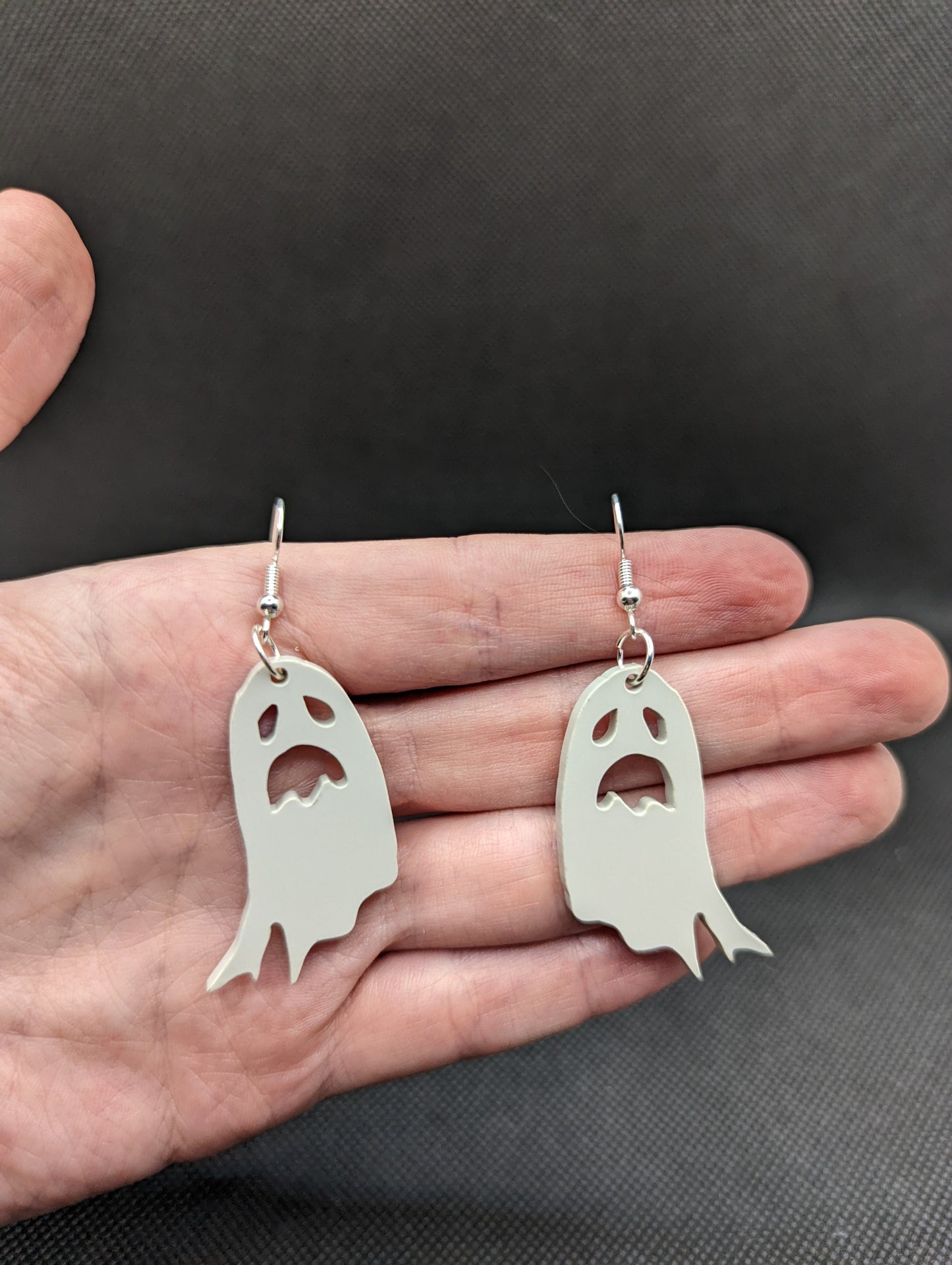 Image of a pair of spooky moaning Ghost shaped earrings on a hand showing the size against a dark background, they are two and a half finger widths length and two wide. They are grey and have silver fixings.