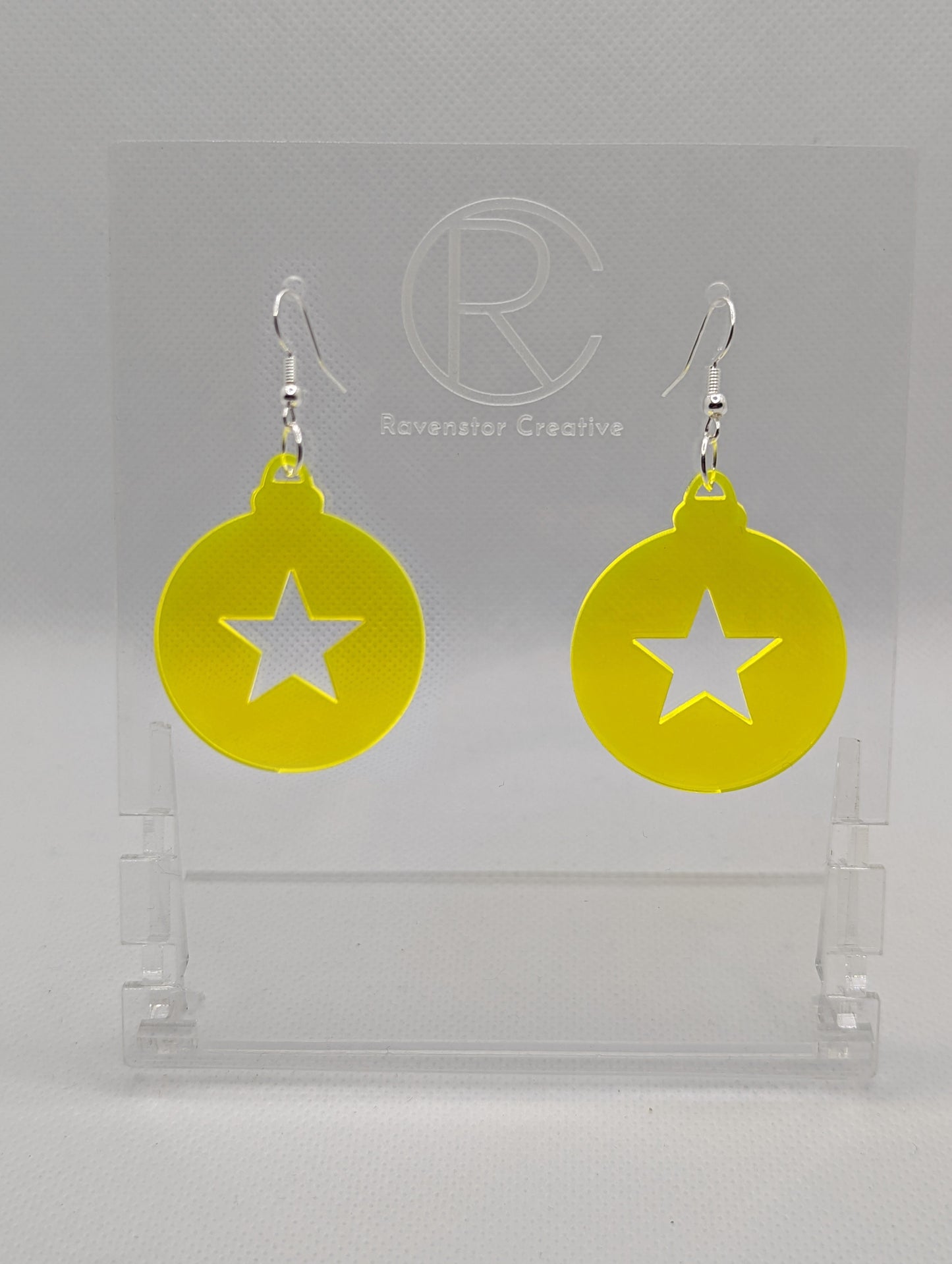 Shows a pair of acrylic earrings that have silver ear wire hook fixings. They are Christmas / Xmas baubles shapes with a star shape cut out the middle. The colour is neon yellow. They are against a white background on see through stands.