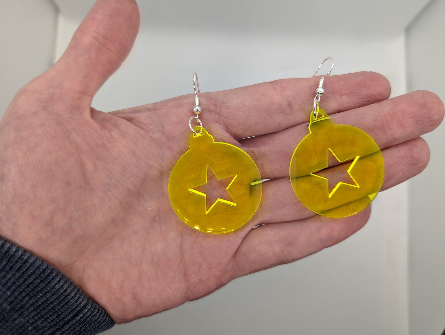 Image of a pair of earrings. They are Christmas / Xmas bauble shapes with a star cut out in Neon Yellow on a hand showing the size against a white background, they are 2 finger widths length the same again wide. They and have silver fixings.