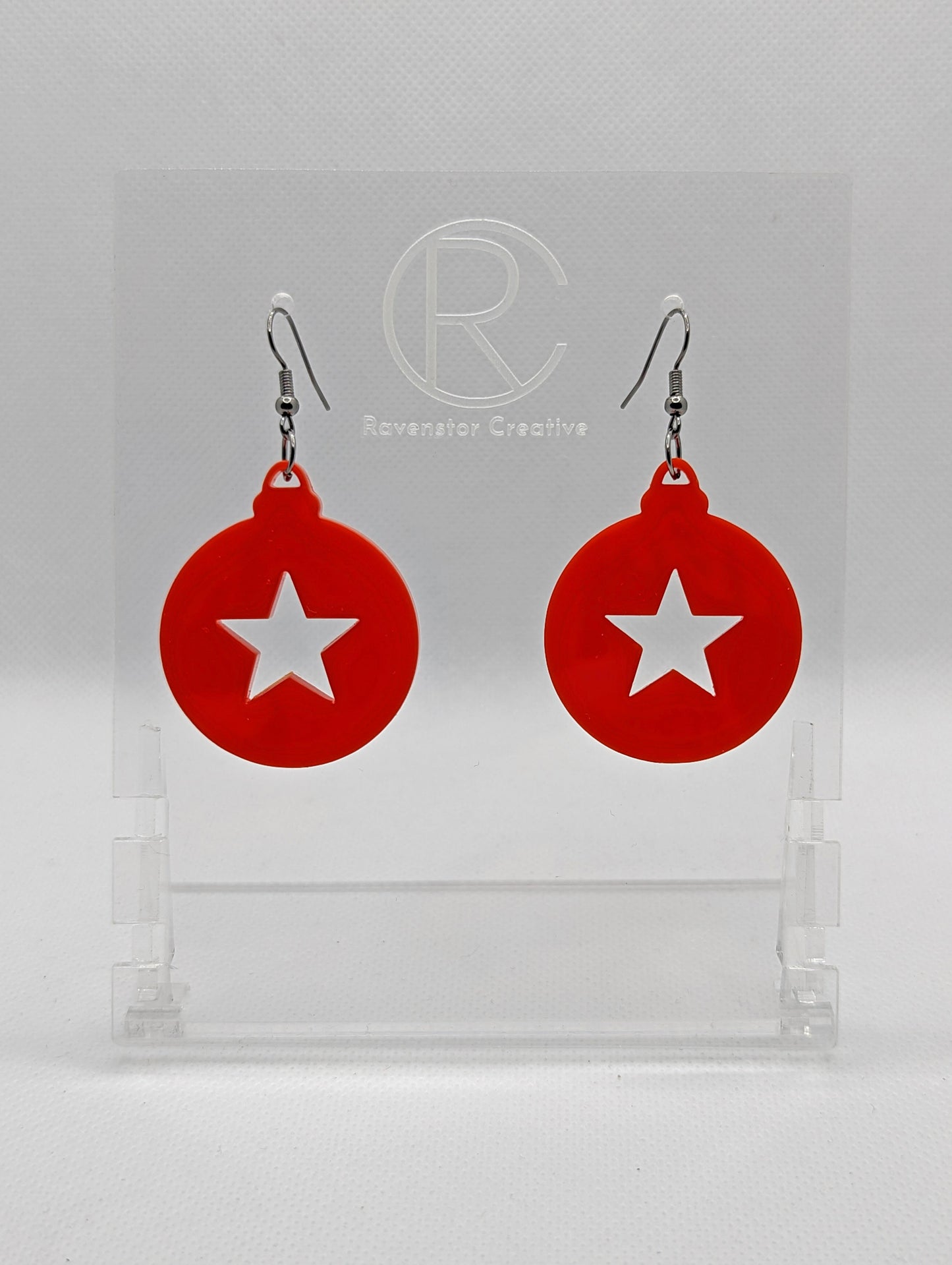 Shows a pair of acrylic earrings that have black ear wire hook fixings. They are Christmas / Xmas baubles shapes with a star shape cut out the middle. The colour is red. They are against a white background on see through stands.