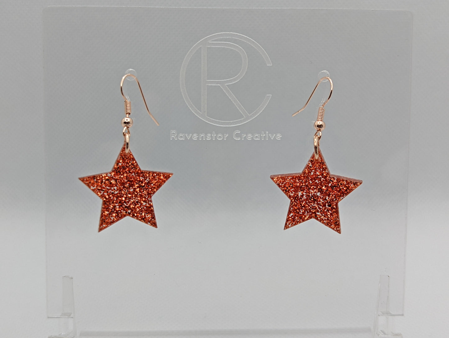 Shows a pair of earrings with rose gold ear wire hook fixings. They are five pointed star shape. The colour of them is copper glitter. They are on a see through stand that has the Ravenstor Creative Logo and its against a white background.
