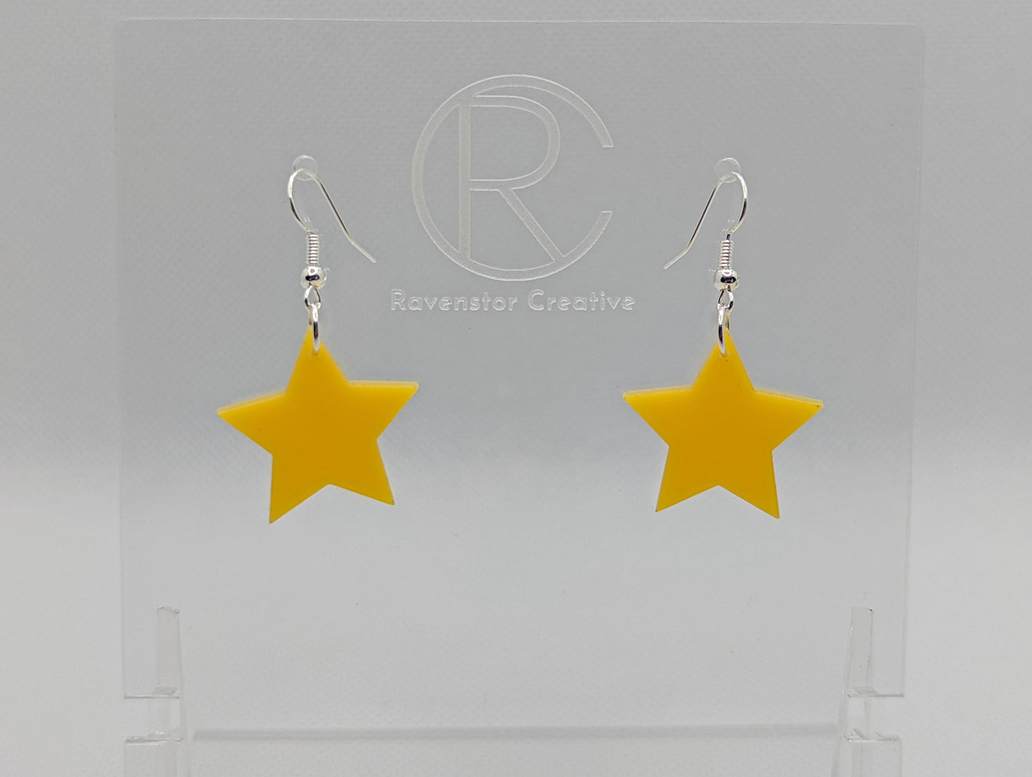 Shows a pair of earrings with silver ear wire hook fixings. They are five pointed star shape. The colour of them is yellow. They are on a see through stand that has the Ravenstor Creative Logo and its against a white background.