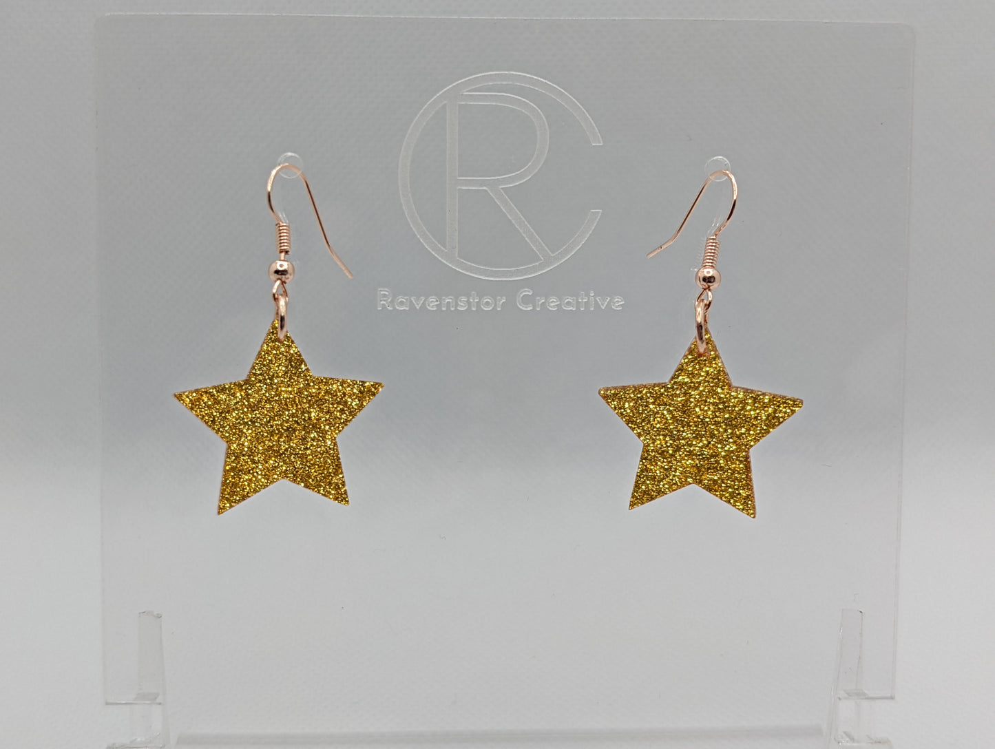 Shows a pair of earrings with rose gold ear wire hook fixings. They are five pointed star shape. The colour of them is gold glitter. They are on a see through stand that has the Ravenstor Creative Logo and its against a white background.