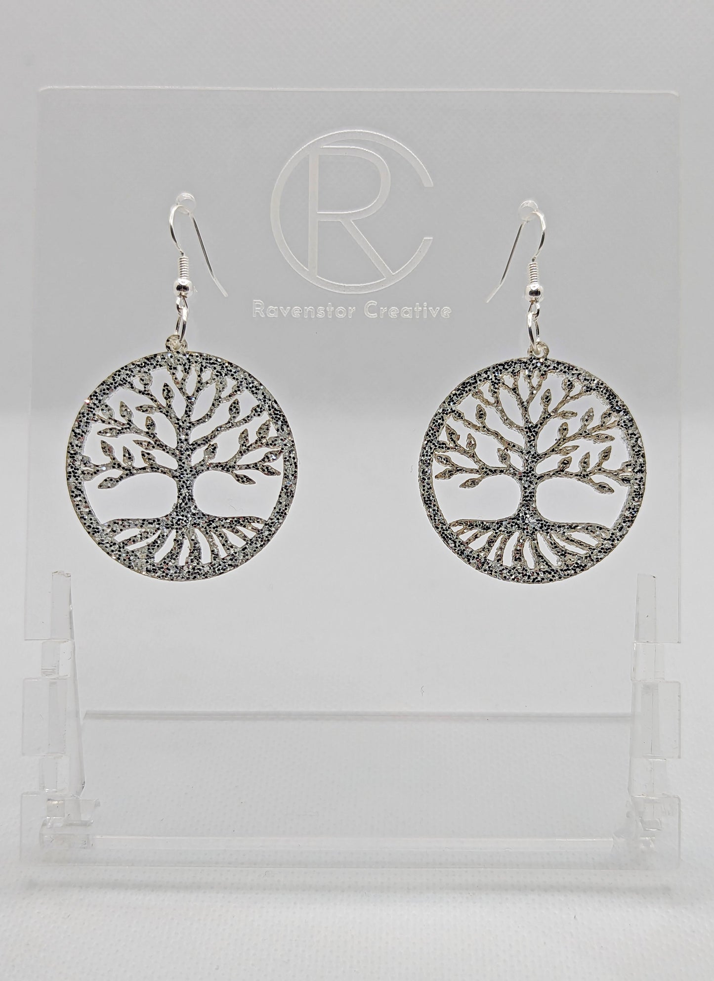 Shows a pair of earrings that have silver ear wire hook fixings. They are circles with the tree of life cut out. They are in Silver Glitter and are against a white background on see through stand with the Ravenstor Creative logo on it.
