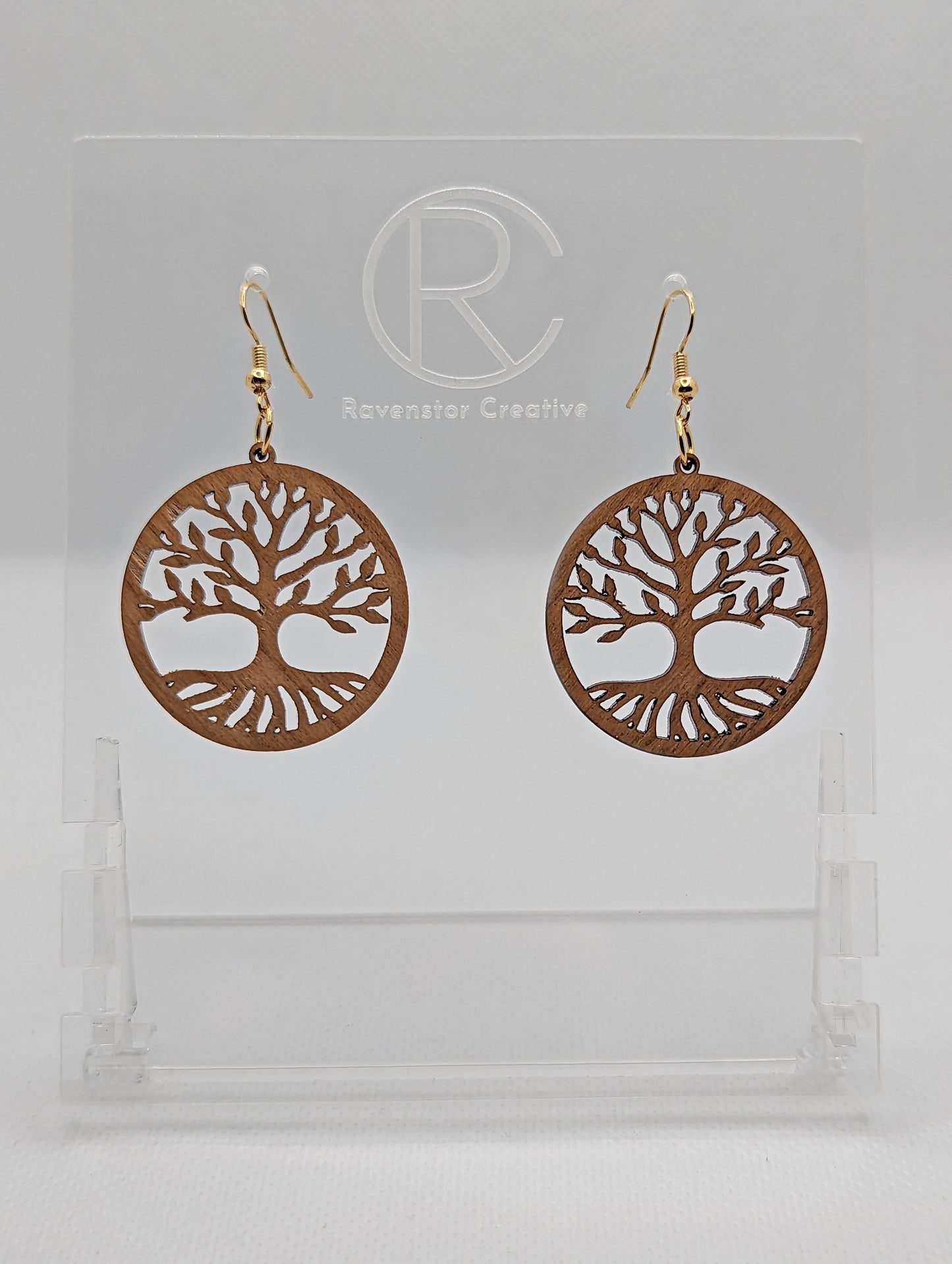 Shows a pair of earrings that have silver ear wire hook fixings. They are circles with the tree of life cut out. They are in Cherry Wood and are against a white background on see through stand with the Ravenstor Creative logo on it.