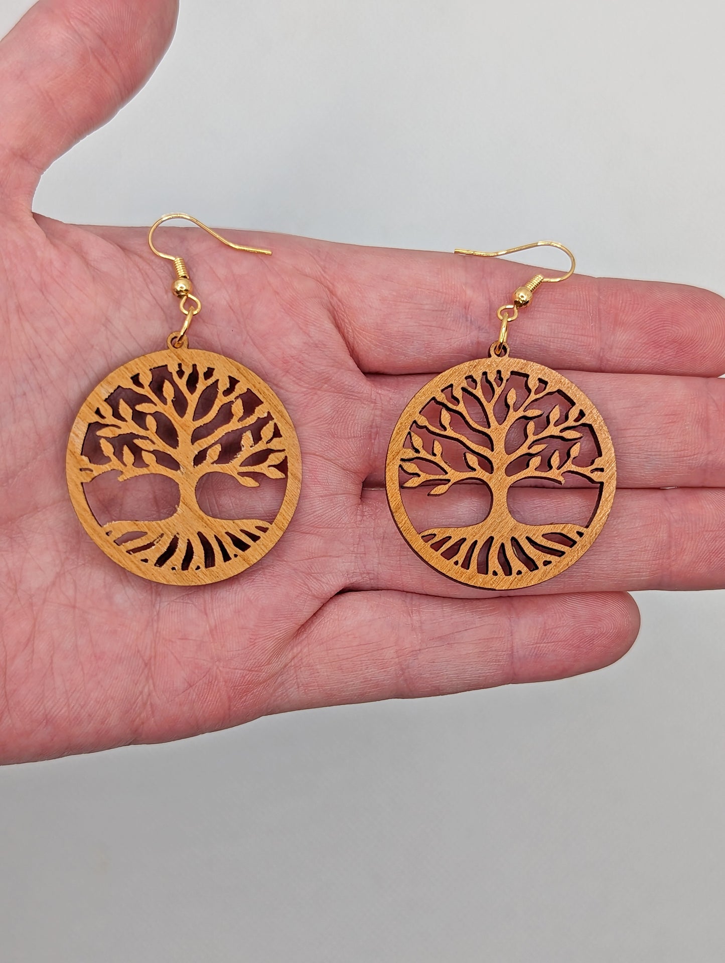 Image of a pair of circle earrings with the tree of life cut out on a hand showing the size against a white background, they are 2 finger widths length the same again wide. They are cherry wood and have gold ear wire hook fixings.