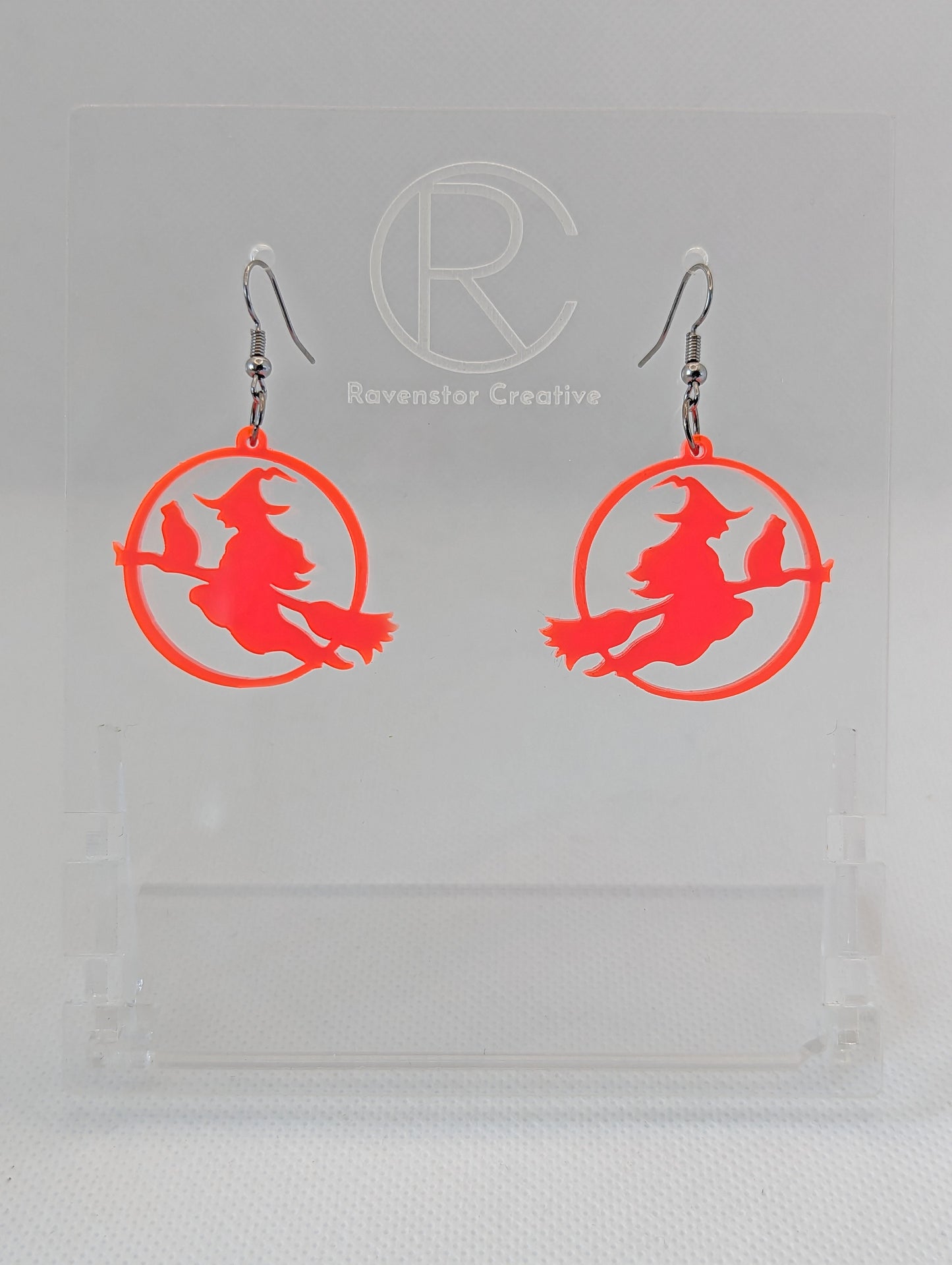 A pair of acrylic earrings with black Ear wire hook fixings on a white background, they are a circle with a scene cut out of a witch on her broom with a cat perched on the broom. The colour is a Neon Pink.