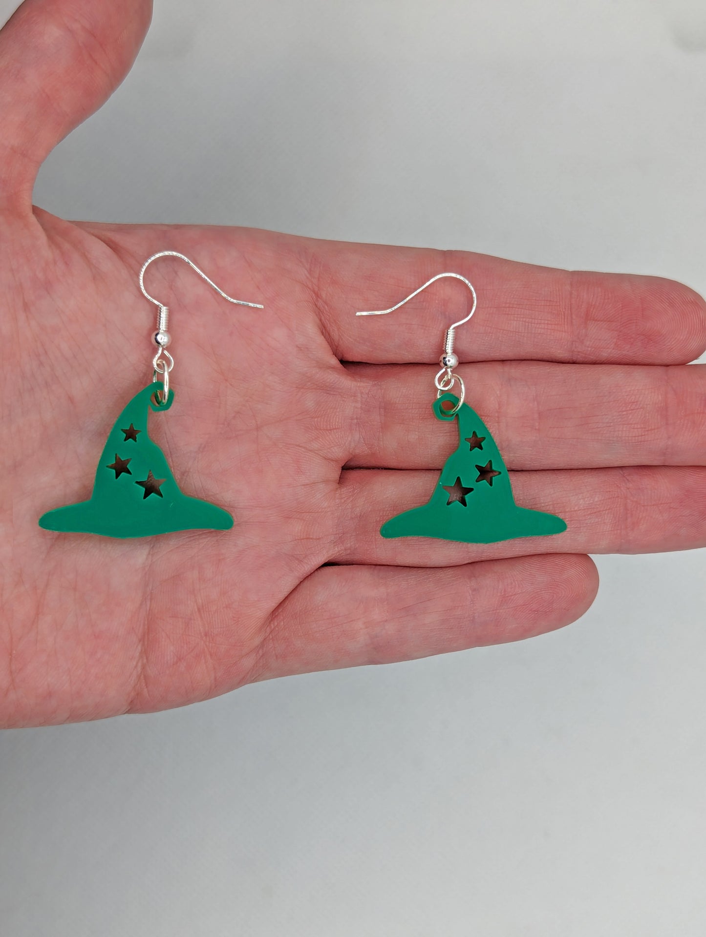 Image of a pair of Halloween Witches Hat shaped earrings with stars cut out on a hand showing the size against a white background, they are two finger widths length and two wide. They are Green and have silver fixings.
