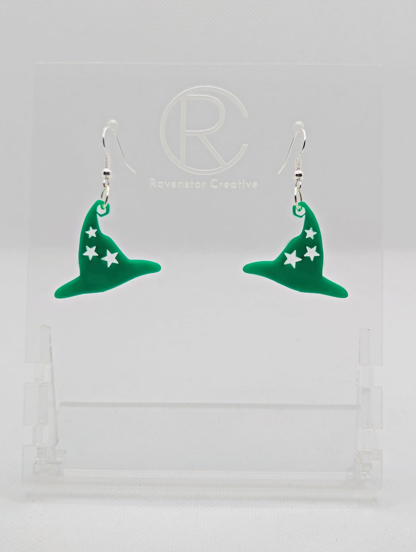 Shows a pair of earrings with silver ear wire hook fixings. They are Halloween Witches hat shapes with stars. The colour of them are Green. They are on a see through stand that has the Ravenstor Creative Logo and its against a white background