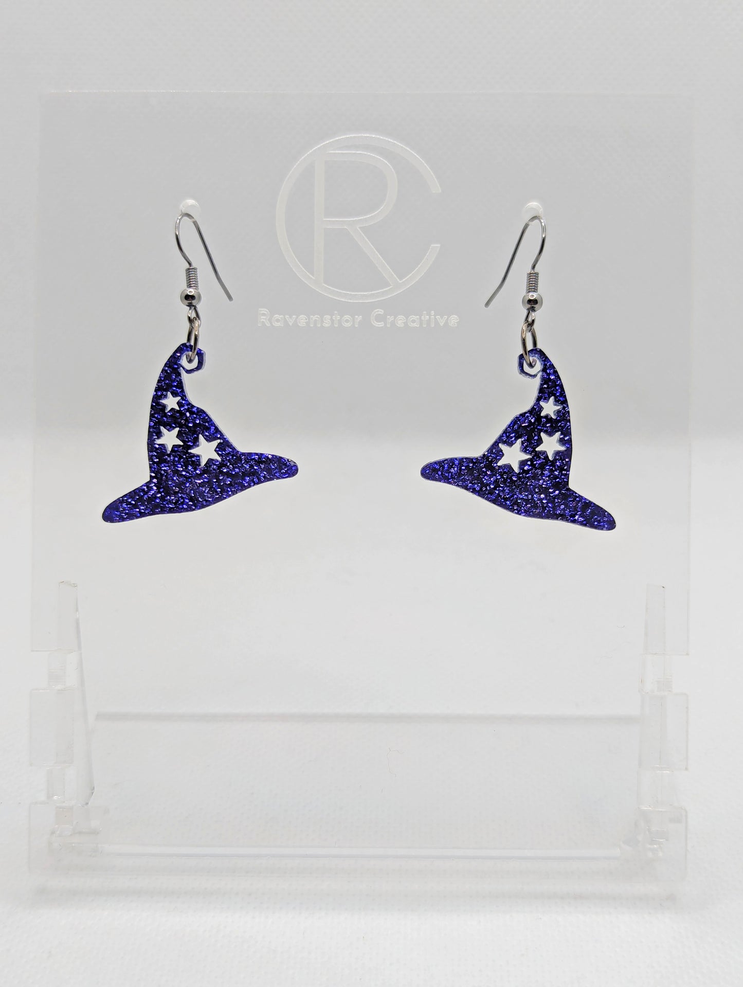 Shows a pair of earrings with black ear wire hook fixings. They are Halloween Witches hat shapes with stars. The colour of them are purple glitter. They on a see through stand that has the Ravenstor Creative Logo and its against a white background