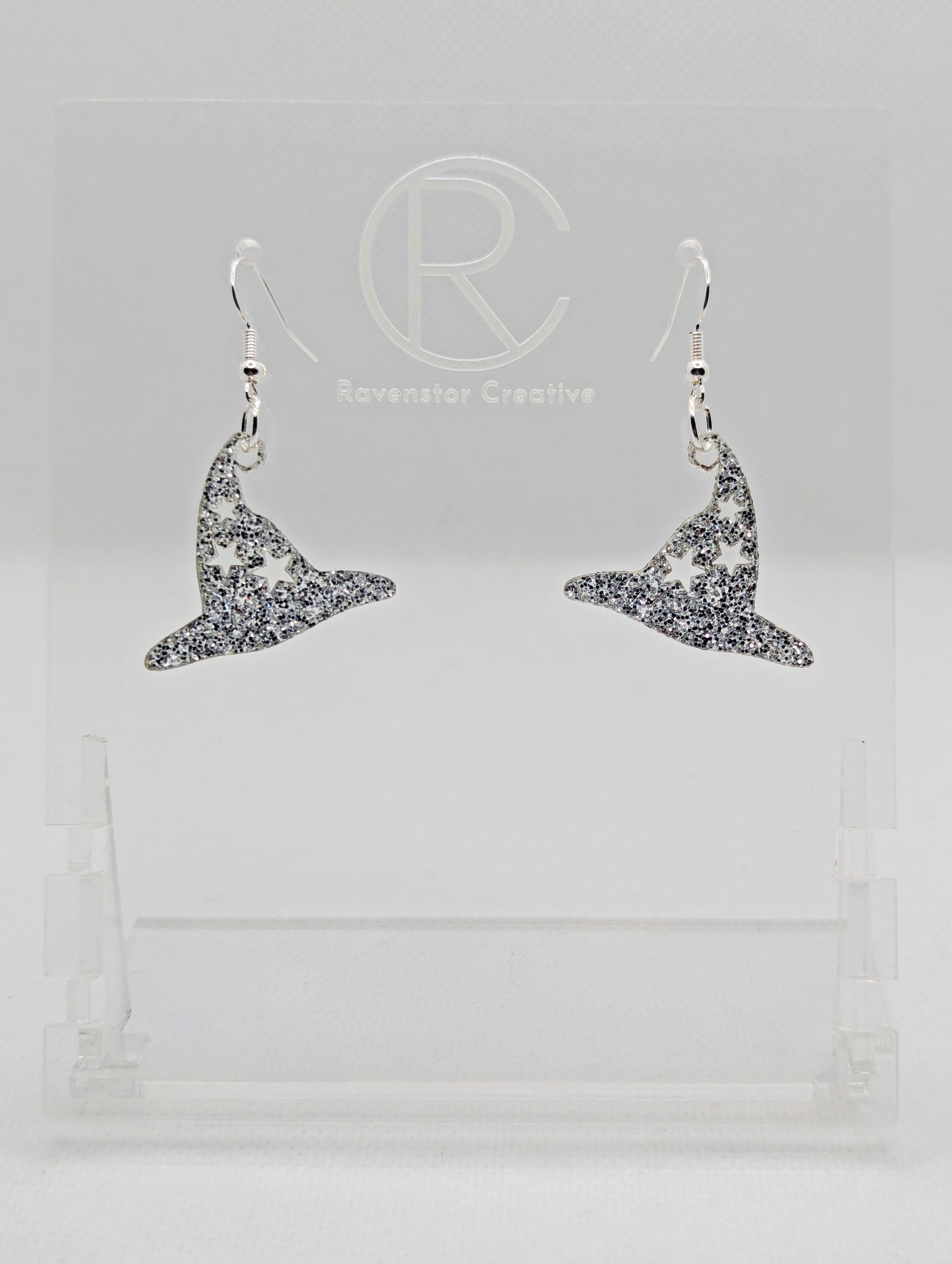 Shows a pair of earrings with silver ear wire hook fixings. They are Halloween Witches hat shapes with stars. The colour of them are silver glitter. They on a see through stand that has the Ravenstor Creative Logo and its against a white background