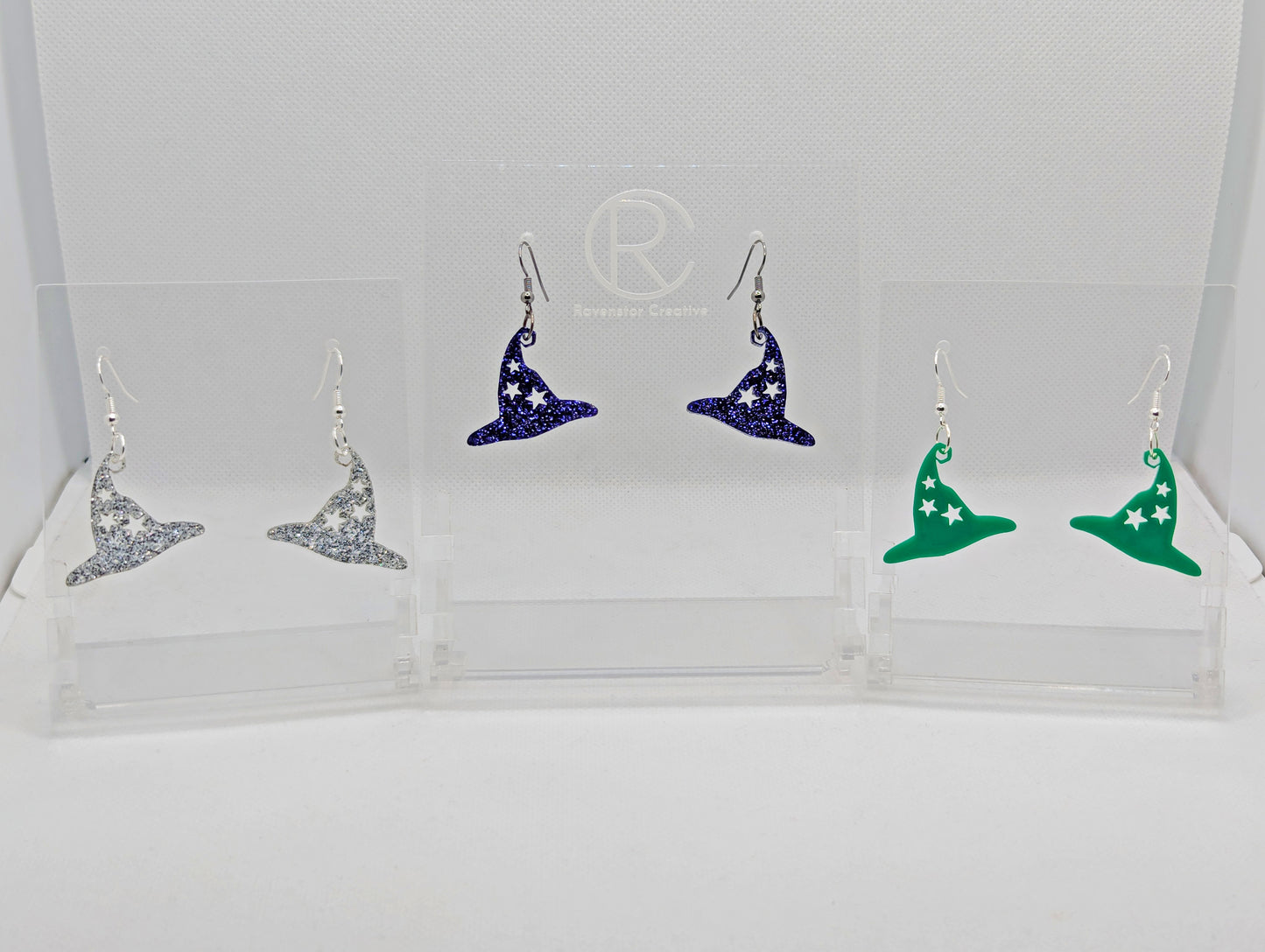 Shows 3 pairs of earrings with ear wire hook fixings. They are Halloween witches hat shapes with 3 starts cut into them. The colours of them are Silver Glitter, Purple Glitter and Green. They are against a white background on clear stands.