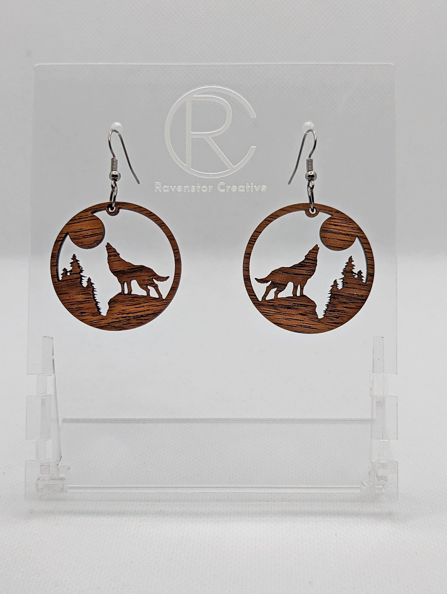Shows a pair of earrings that have black ear wire hook fixings. They are circles with a wolf howling at the moon cut out. They are in walnut wood and are against a white background on see through stand with the Ravenstor Creative logo on it.