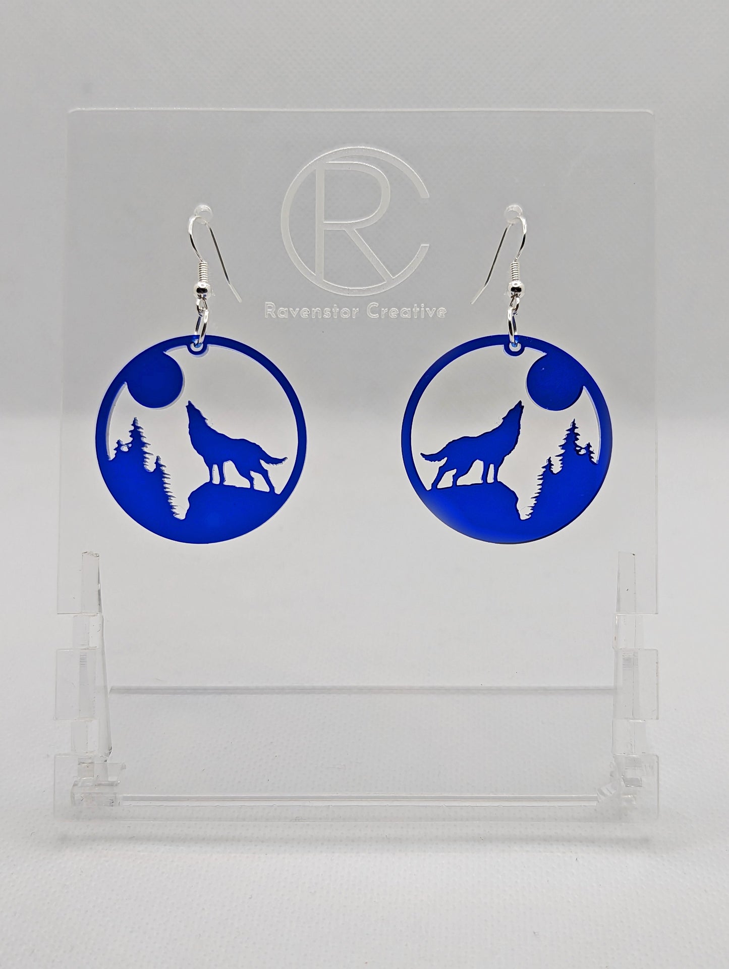 Shows a pair of earrings that have silver ear wire hook fixings. They are circles with a wolf howling at the moon cut out. They are in translucent blue and are against a white background on see through stand with the Ravenstor Creative logo on it.