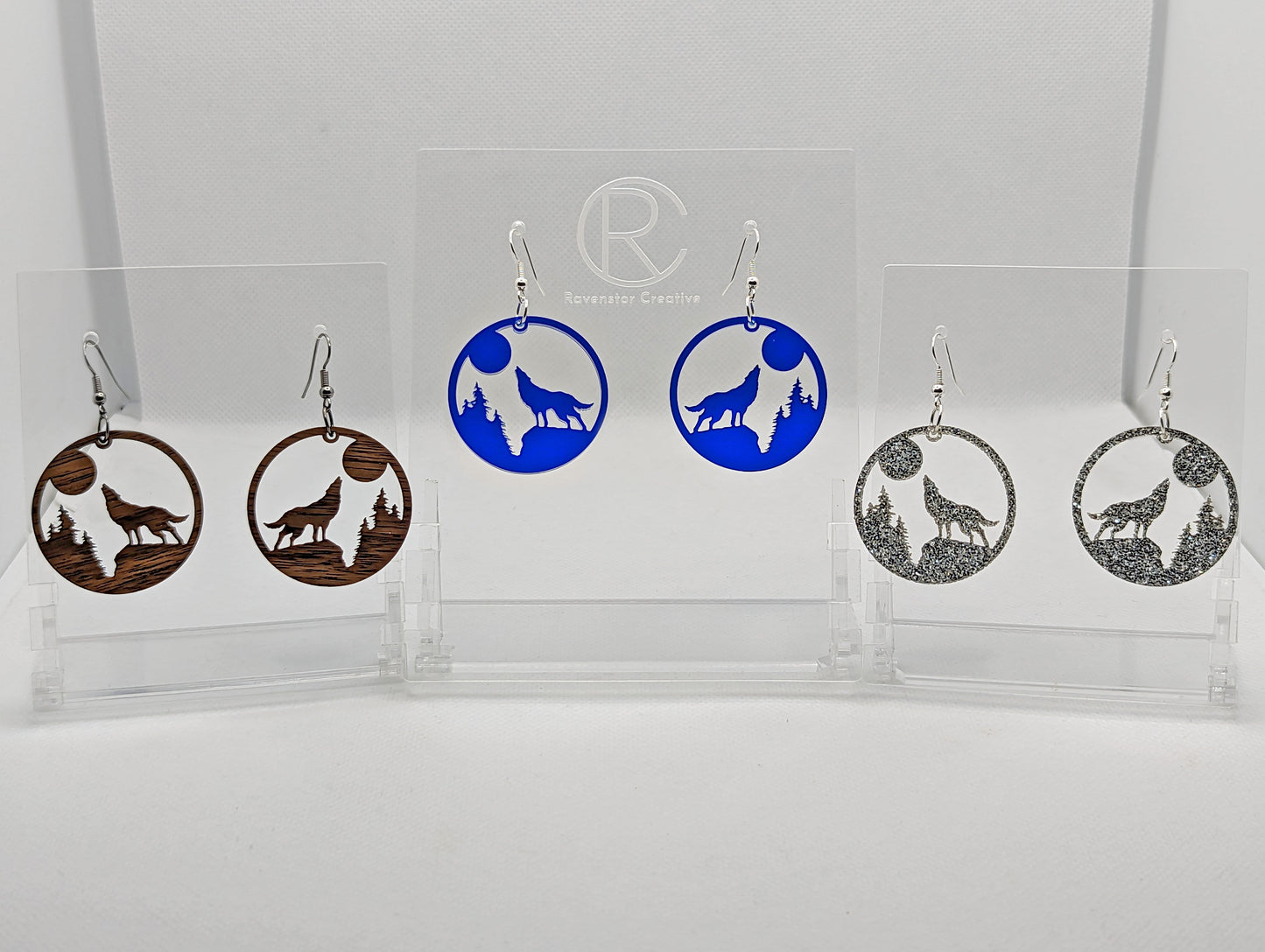 Shows three pairs of earrings that have ear wire hook fixings. They are a circle with a wolf howling at the moon cut out. The colours are walnut wood, translucent blue and Silver Glitter. They are against a white background on see through stands.