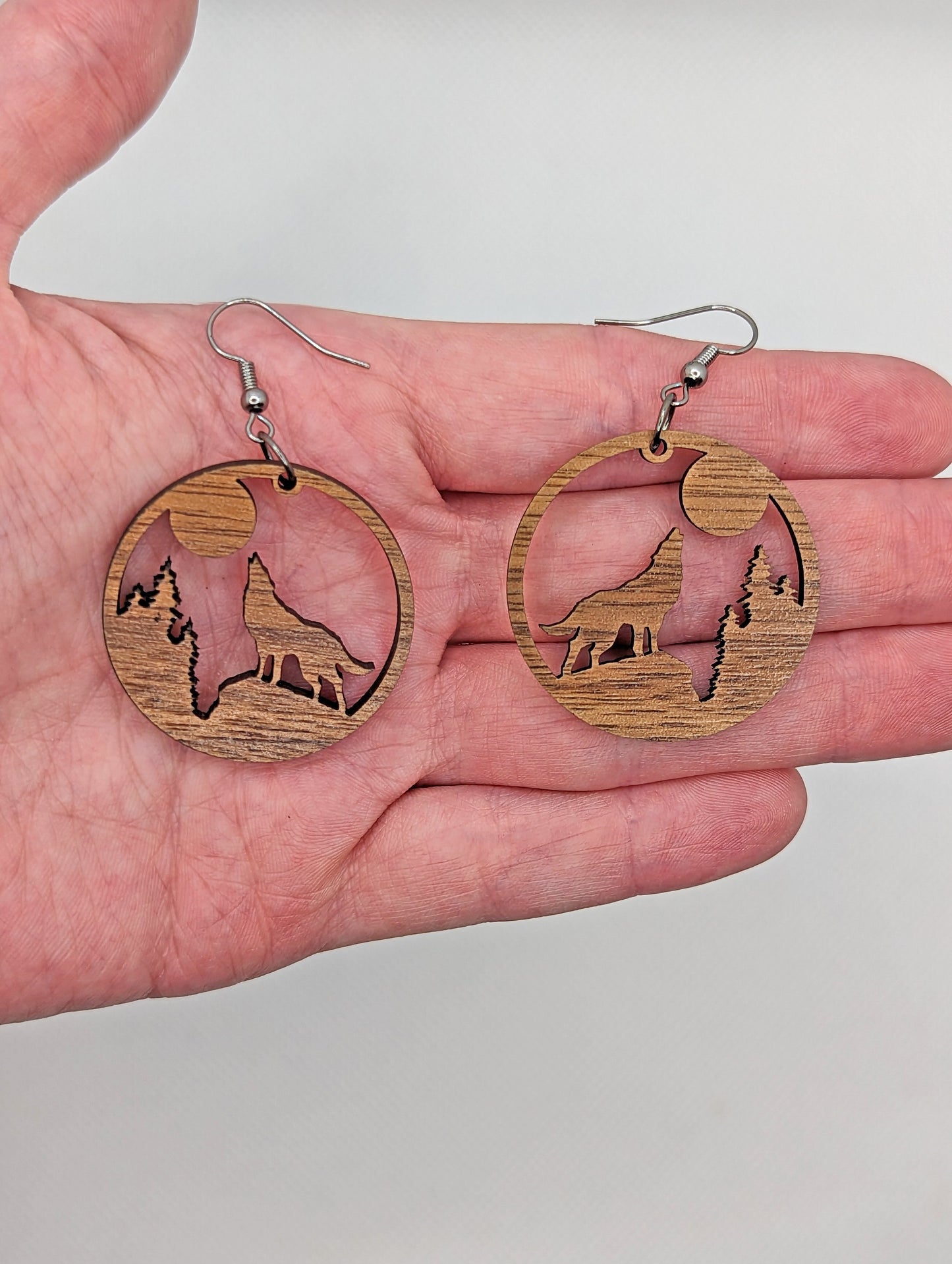 Image of a pair of circle earrings a wolf howling at the moon cut out, on a hand showing the size against a white background, they are 2 finger widths length the same again wide. They are walnut wood and have black fixings.
