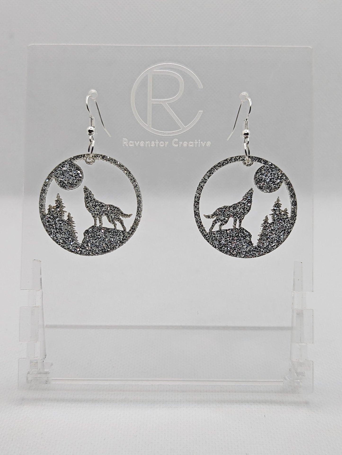 Shows a pair of earrings that have silver ear wire hook fixings. They are circles with a wolf howling at the moon cut out. They are in Silver Glitter and are against a white background on see through stand with the Ravenstor Creative logo on it.