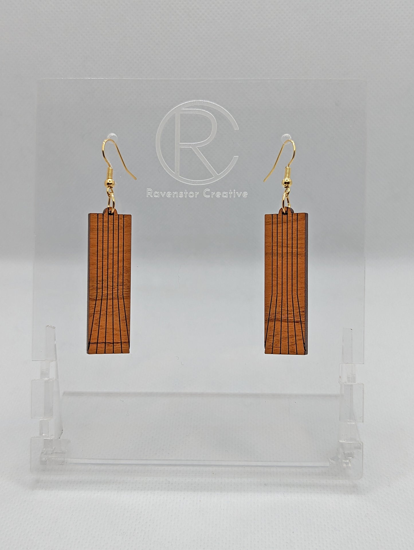 A pair of earrings made in Cherry Wood with gold ear wire hook fixings placed on a clear stand on a white background. They are a slim rectangle with vertical lines running from the top to the bottom but they fan out slightly just below half way down.
