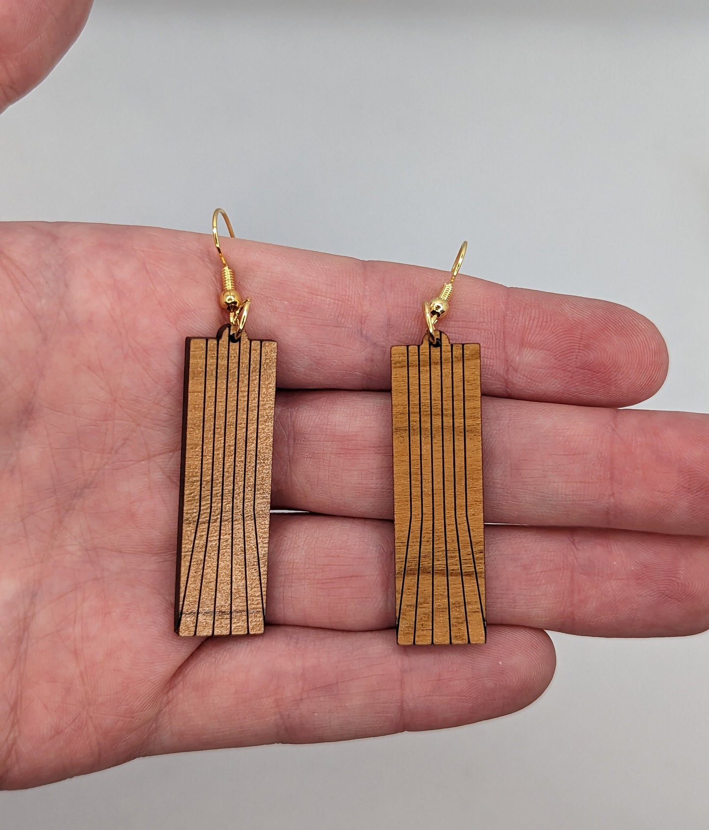 A pair of earrings on a hand showing the size against a white background, they are two and a half finger widths in length and one wide . They are a slim rectangle with vertical lines running from the top to the bottom that fan out.