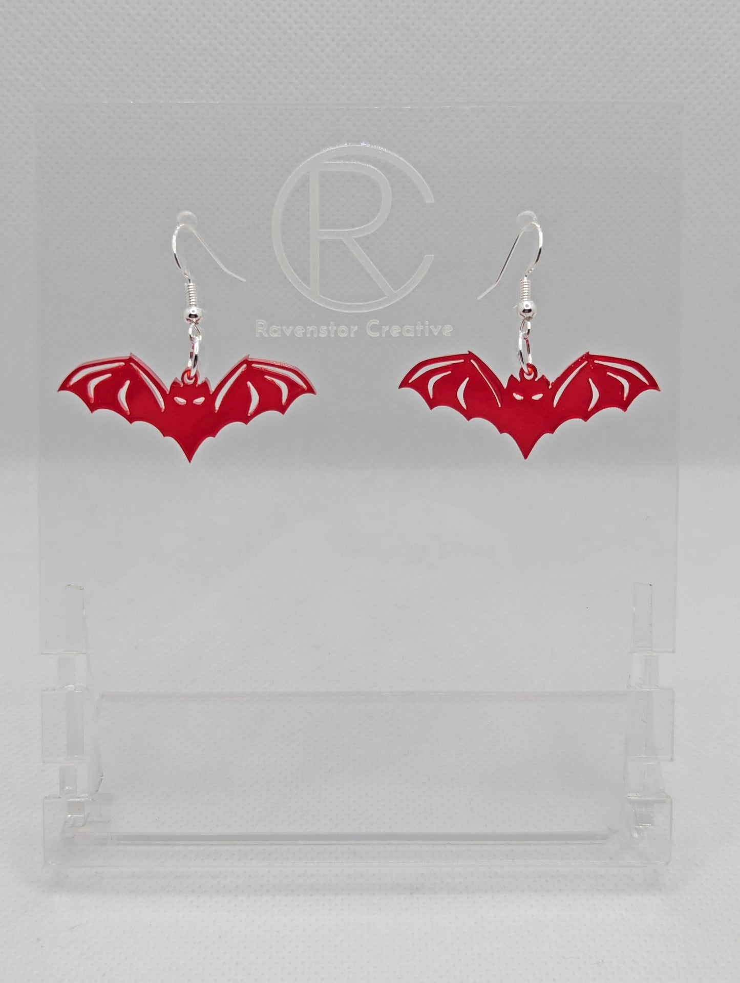 Shows a pair of earrings with silver ear wire hook fixings. They are Halloween Bat shapes. The colour of them is translucent red. They are on a see through stand that has the Ravenstor Creative Logo and its against a white background.