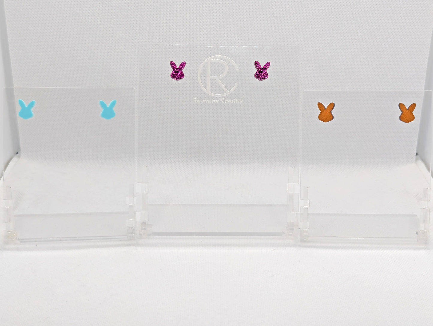 Shows three pairs of small studded earrings that are Bunny / Rabbit Playboy heads. The colours are Spearmint, Pink Glitter and Cherry Wood. They are against a white background on a clear stand with the Ravenstor Creative logo on it.