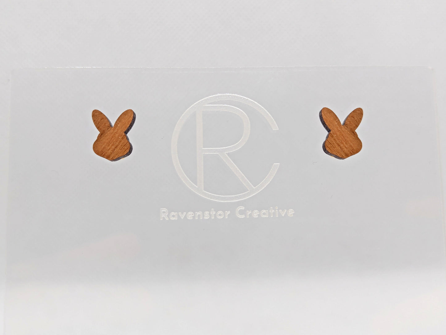 Shows a pair of small studded wooden earrings that are Bunny / Rabbit heads. They are Cherry Wood in colour. They are against a white background on see through stand with the ravenstor creative logo on.