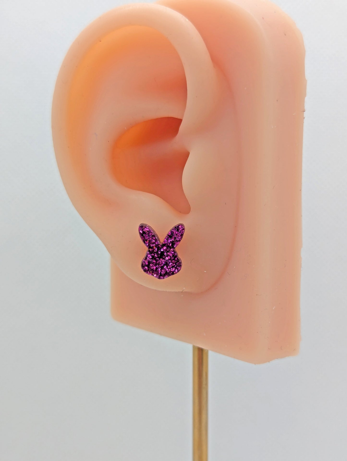 Shows a pair of small studded acrylic earrings that are Bunny / Rabbit Playboy Heads. They are Pink Glitter in colour. They are on an ear to show the size.