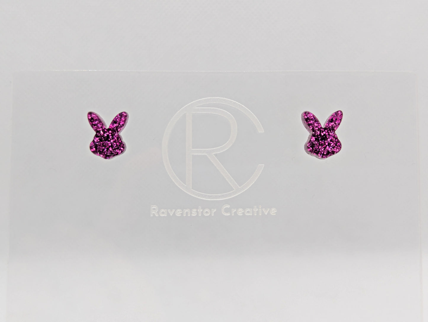 Shows a pair of small studded acrylic earrings that are Bunny / Rabbit heads. They are Pink Glitter in colour. They are against a white background on see through stand with the ravenstor creative logo on.