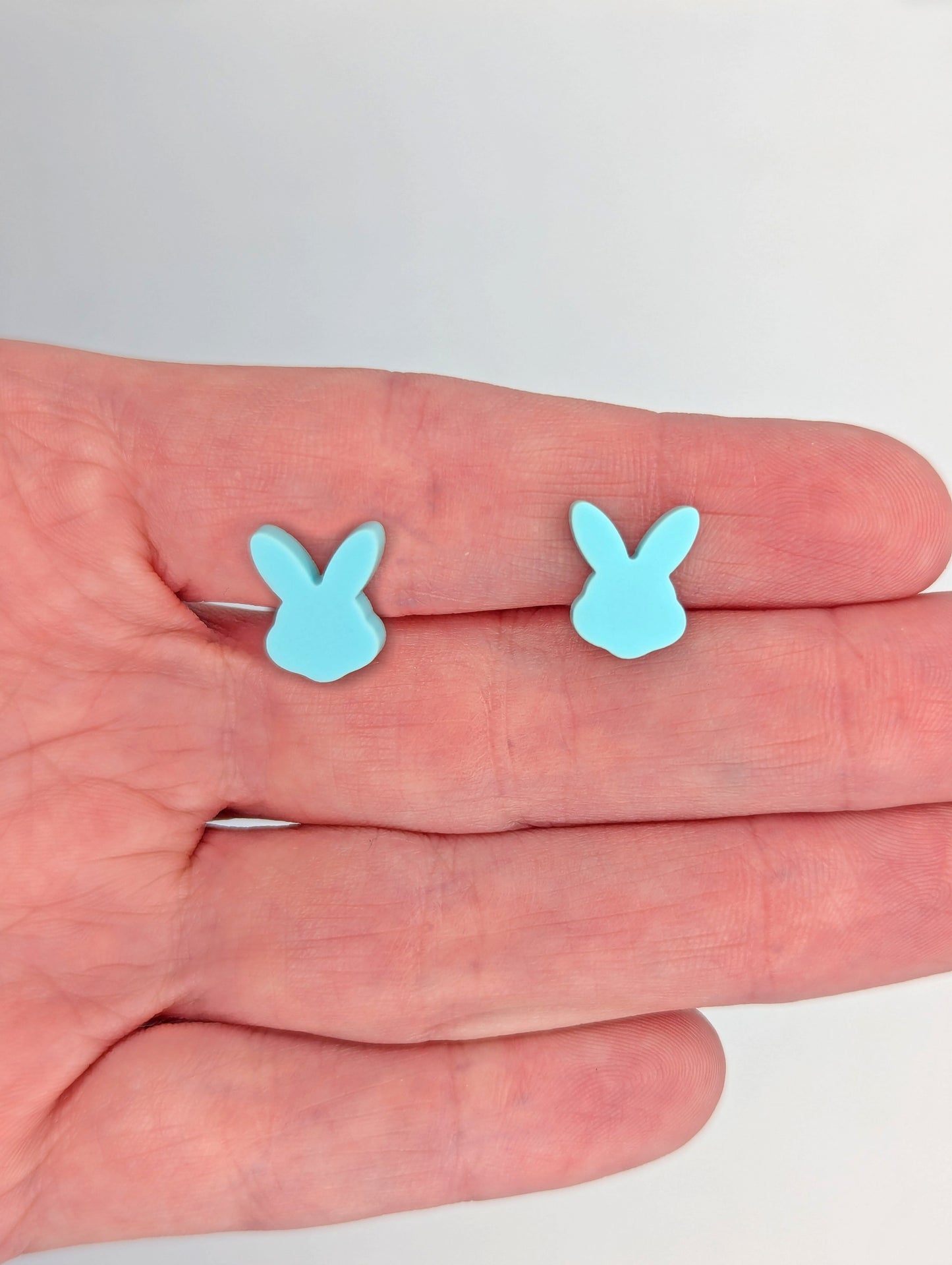 Shows a pair of small studded acrylic earrings that are Bunny / Rabbit heads. The colour is Spearmint. They are against a hand showing the size of one finger width in length and slightly smaller width.