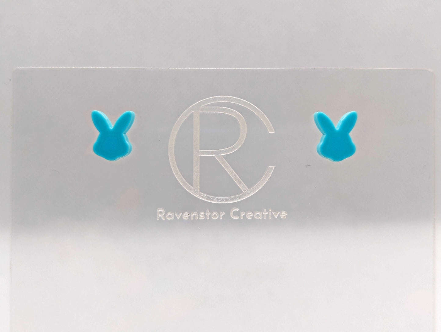 Shows a pair of small studded acrylic earrings that are Bunny / Rabbit heads. They are Spearmint in colour. They are against a white background on see through stand with the ravenstor creative logo on.