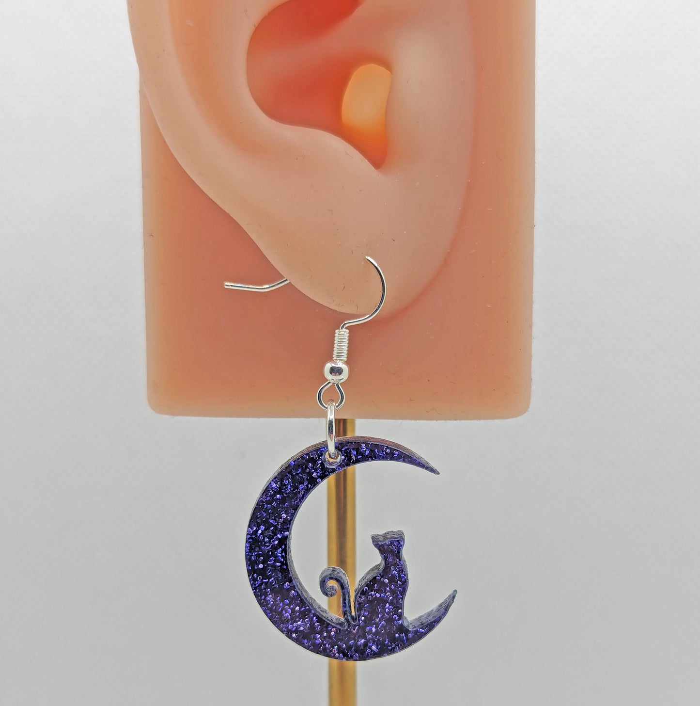 Image of a pair of earrings with a cat sitting in a crescent moon, on an ear showing the size against a white background. They are purple glitter and have silver fixings.