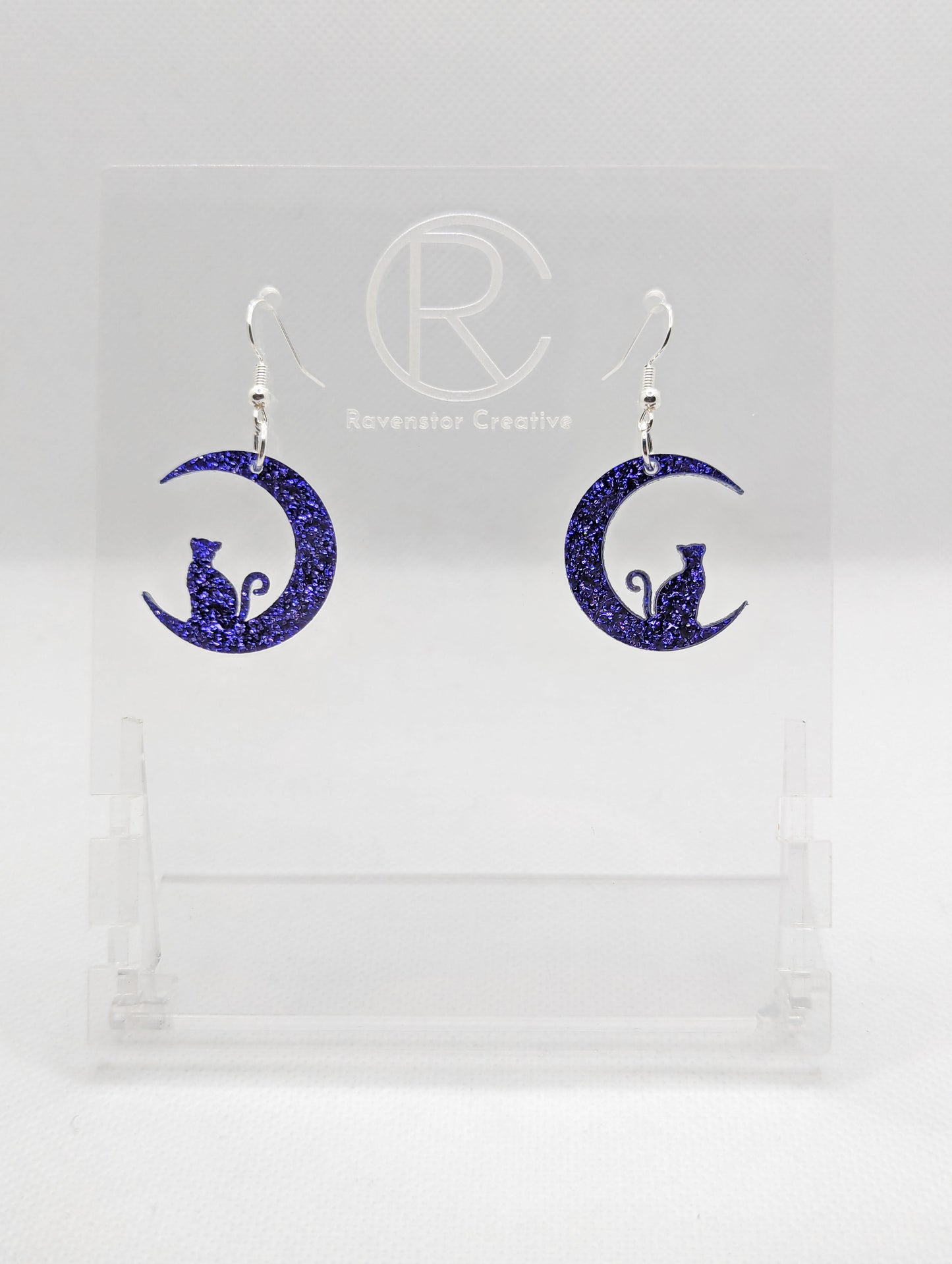 Shows a pair of acrylic earrings that have silver ear wire hook fixings. They are a cat sitting in a crescent moon. They are in Purple Glitter and are against a white background on see through stand with the Ravenstor Creative logo on it.