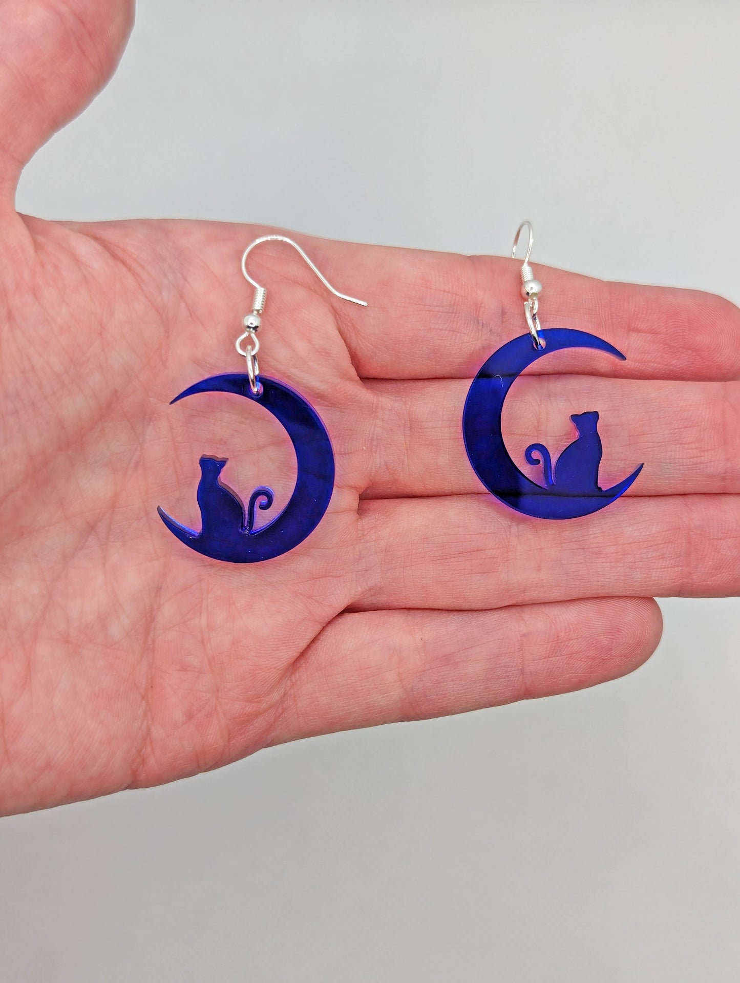 Image of a pair of earrings with a cat sitting in a crescent moon, on a hand showing the size against a white background, they are 2 finger widths length the same again wide. They are translucent blue and have silver fixings.