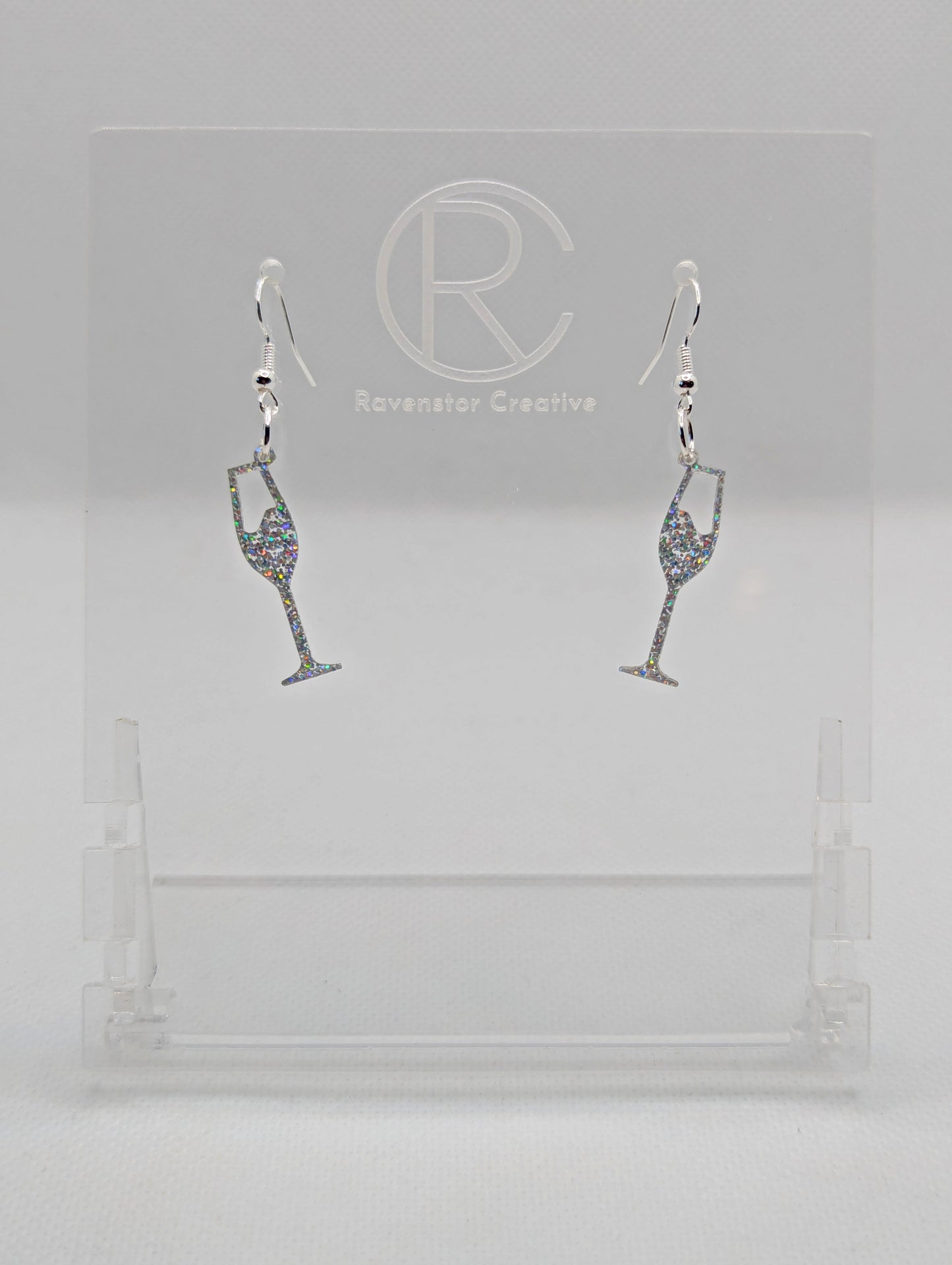 An image of a pair of Champaign Glass Earrings with Silver ear wire hook fixings. These are ideal for a Hen Do or Bachelorette Party. The colour is Holographic Glitter. They are on clear stands against a white background.