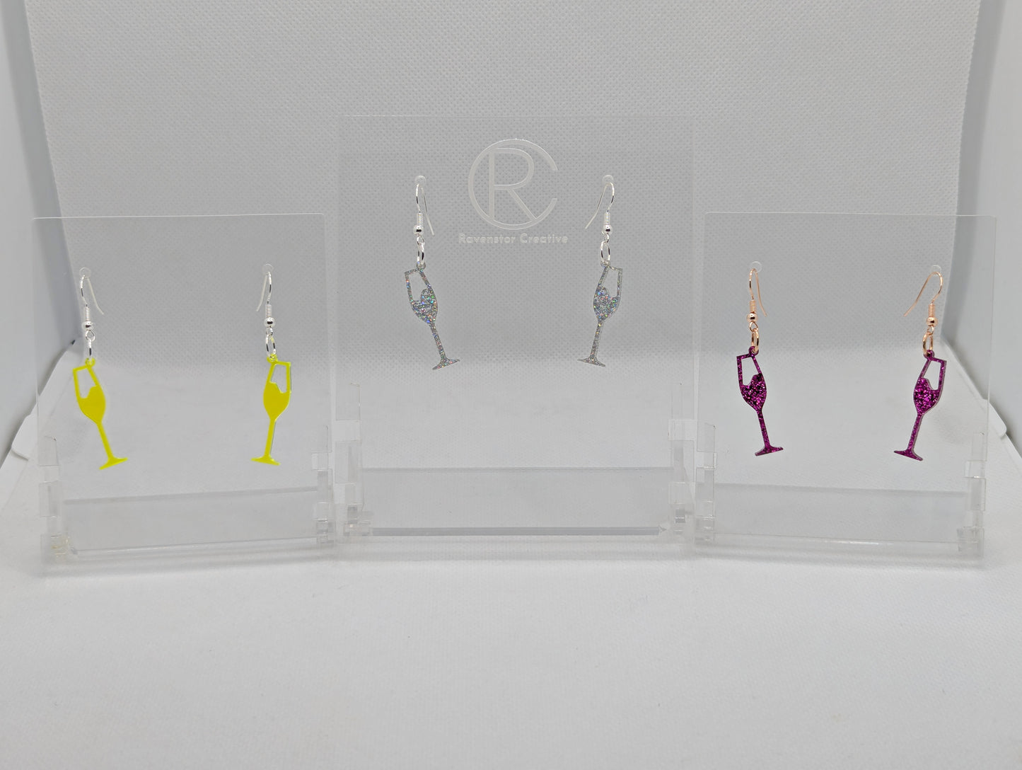 An image of three pairs of Champaign Glass Earrings with ear wire hook fixings. These are ideal for a Hen Do or Bachelorette Party. The colours are Neon Orange, Holographic Glitter and Pink Glitter. The are on clear stands against a white background.