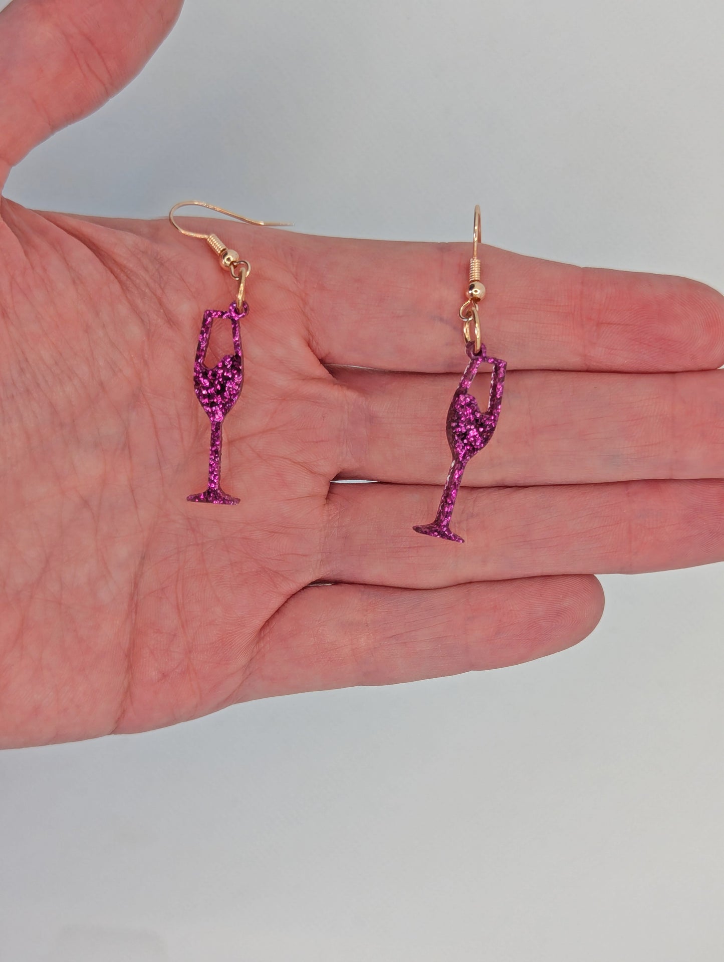 An image of a pair of Champaign Glass Earrings with Rose Gold ear wire hook fixings. These are ideal for a Hen Do or Bachelorette Party. They are against a hand to show the size of two finger widths in length and half a width wide.